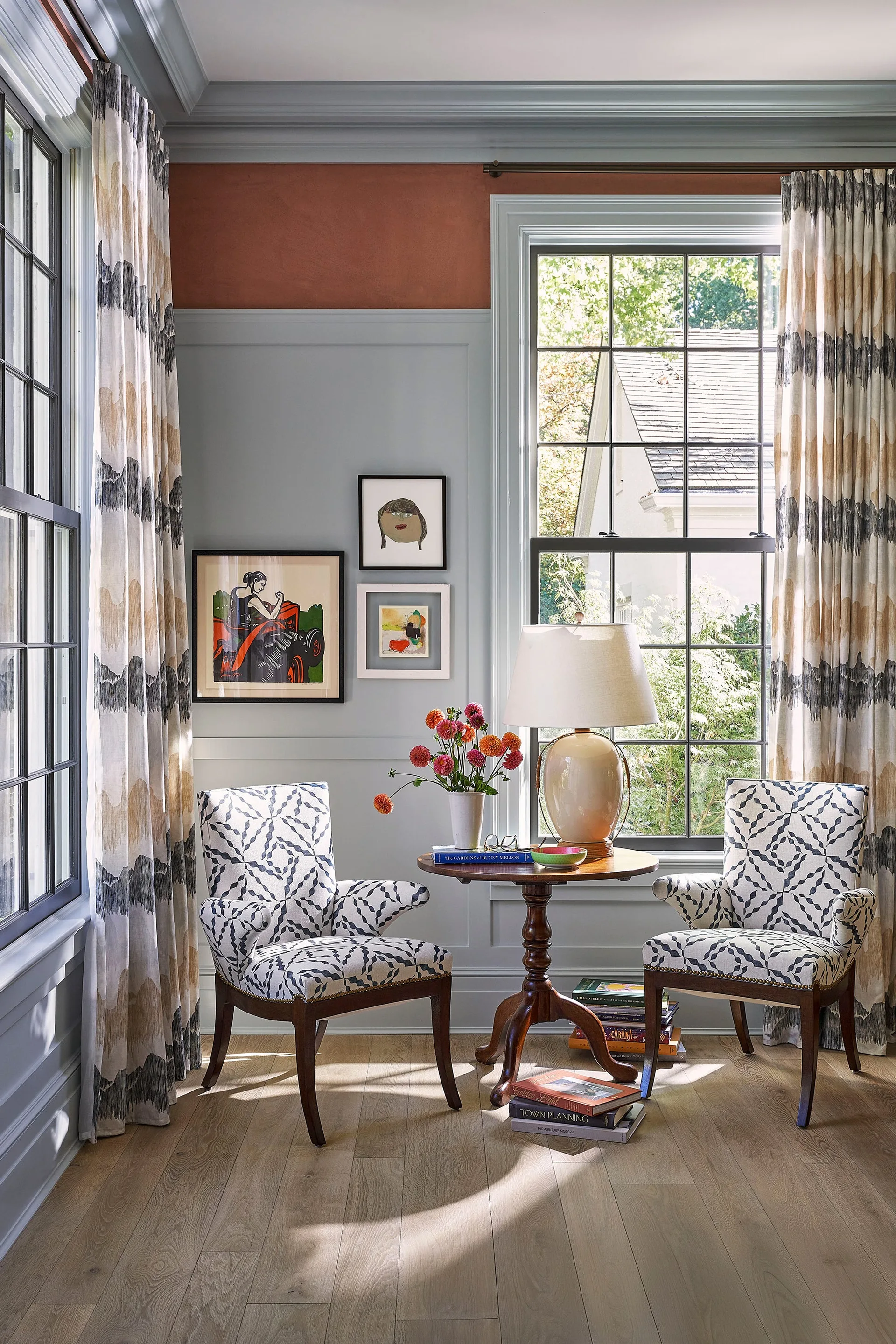 Two patterned armchairs flank a dark wood pedestal table in front of a window with light blue wainscoting.