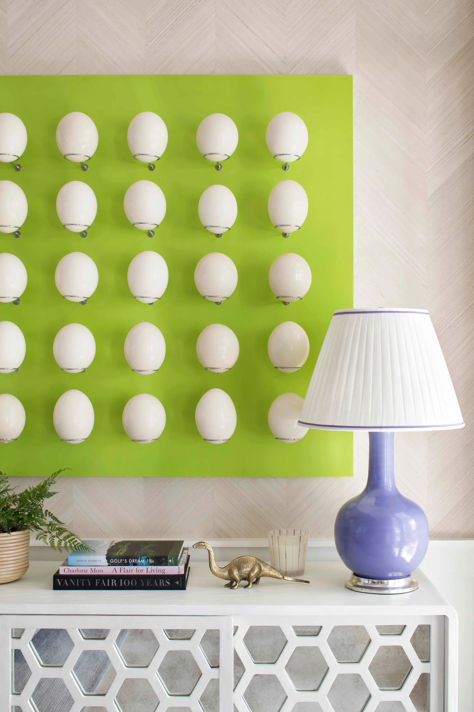 A white console table with honeycomb doors features a purple lamp and books beneath a bright green wall panel decorated with rows of white spheres.