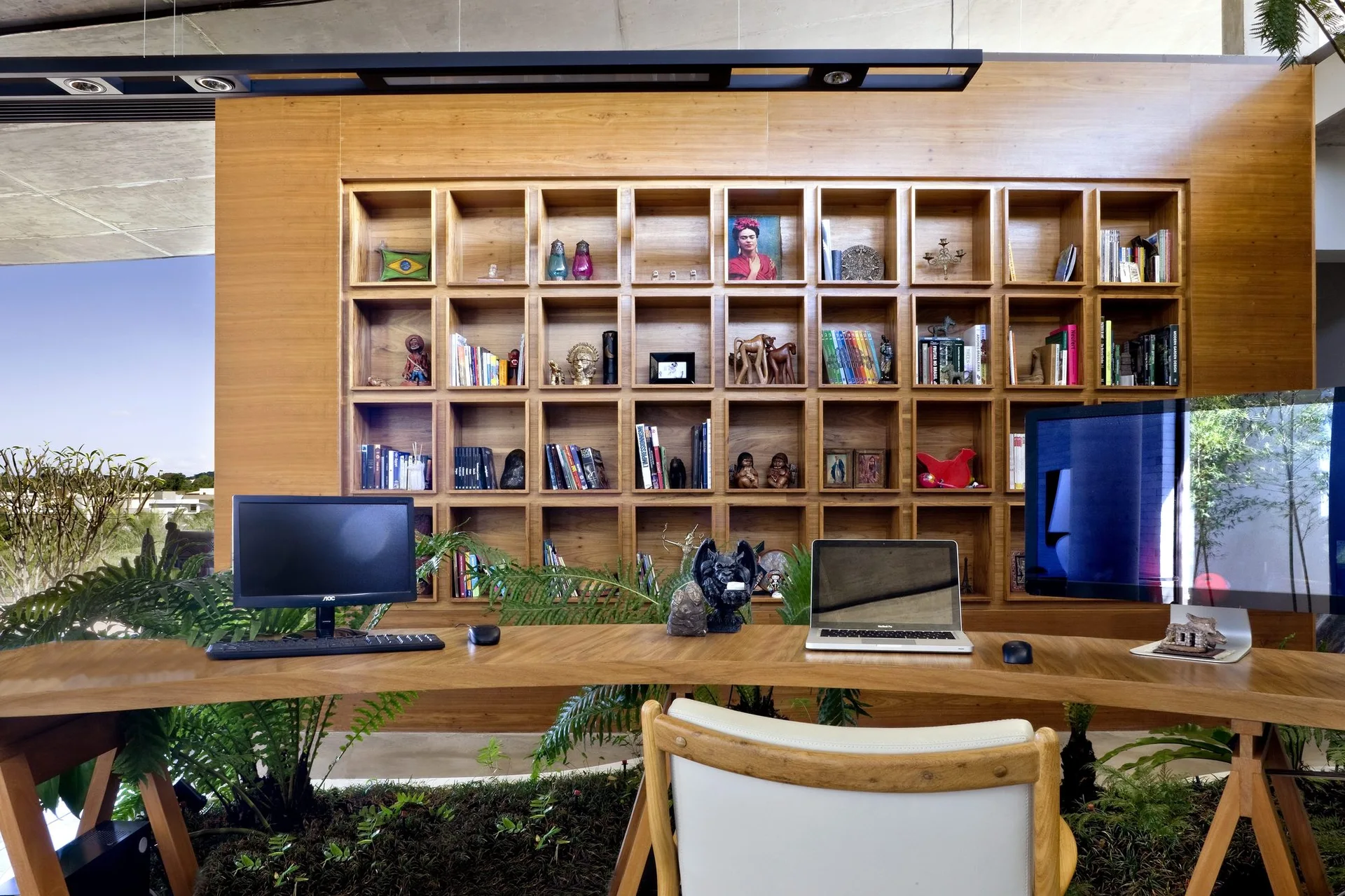 A modern home office features a curved wooden desk, a built-in grid bookshelf wall, and green plants integrated into the floor.