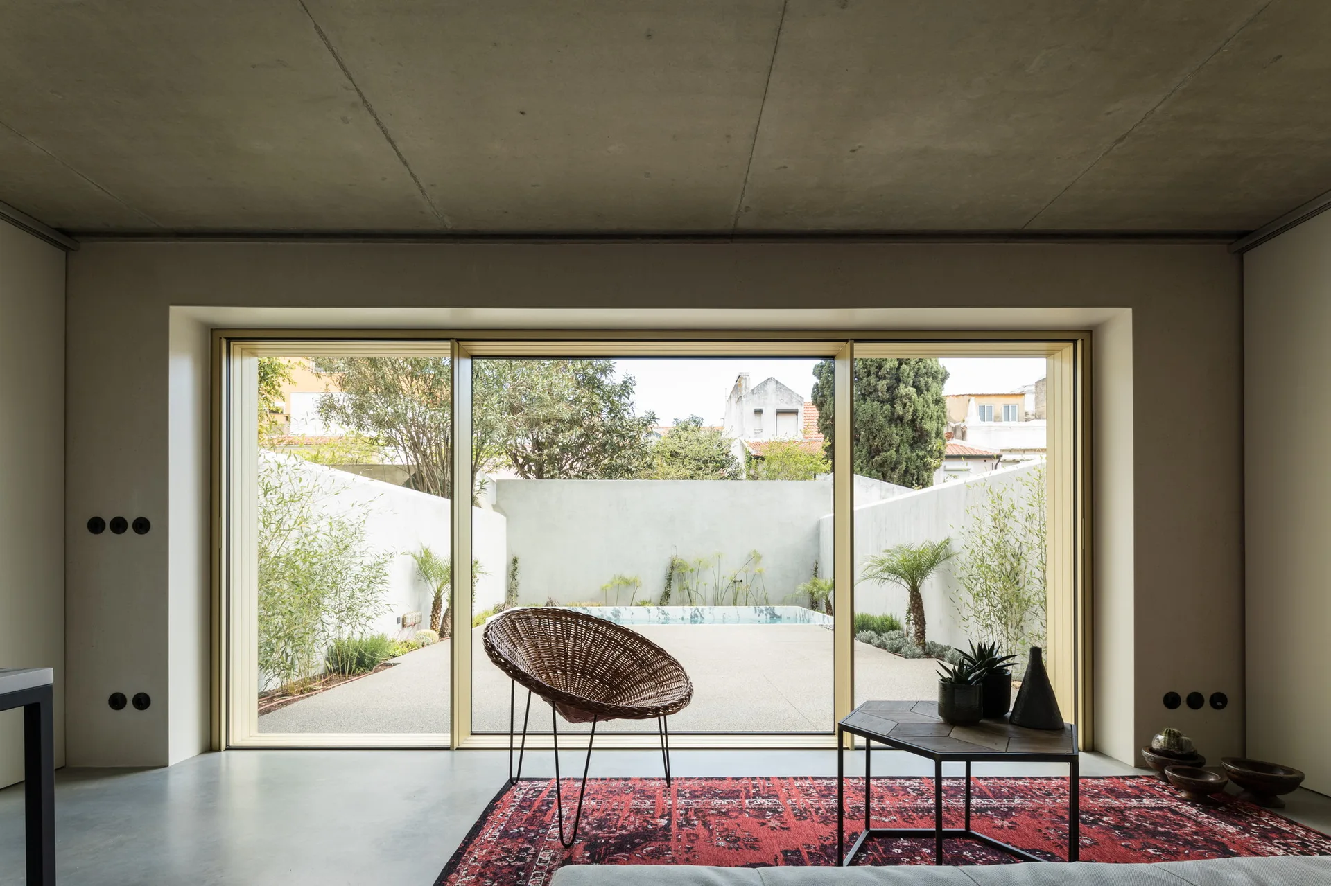A minimalist bedroom features an exposed concrete ceiling, a large sliding glass door opening to a patio, a red patterned rug, and a wicker chair.