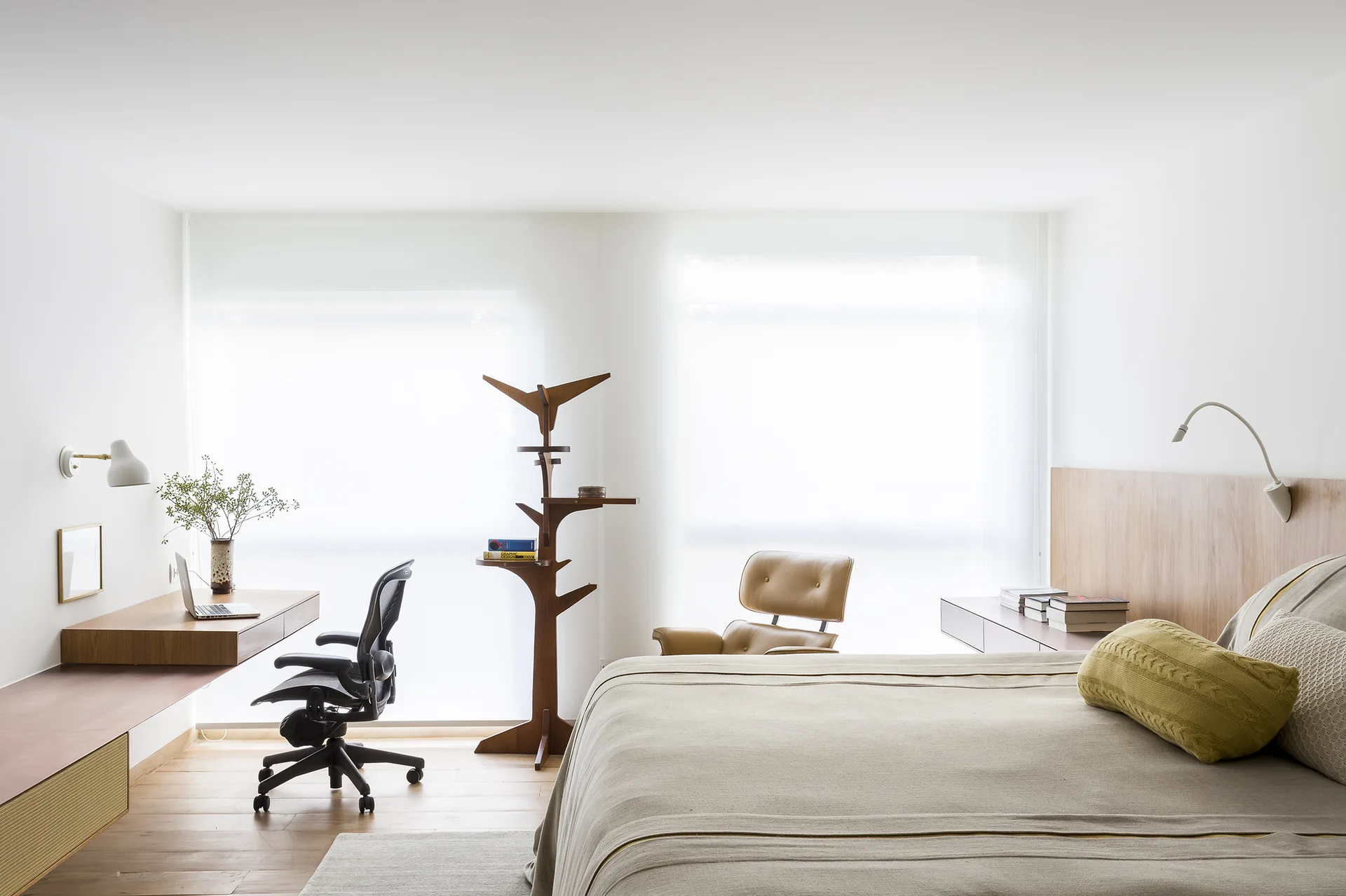 A minimalist bedroom with a floating wooden desk, black office chair, bed with integrated wood headboard, and large windows with sheer shades.