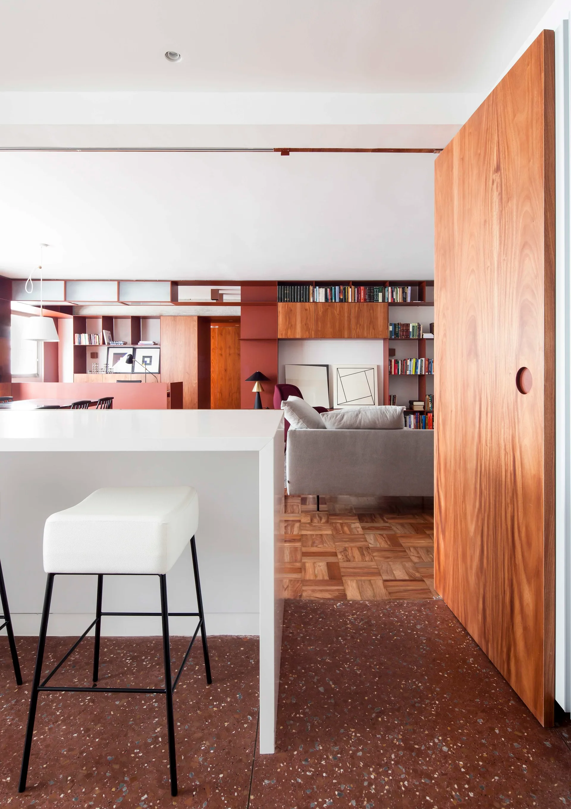 A modern interior features a white kitchen island, a grey living room sofa, a warm wood sliding door, and red built-in shelving.