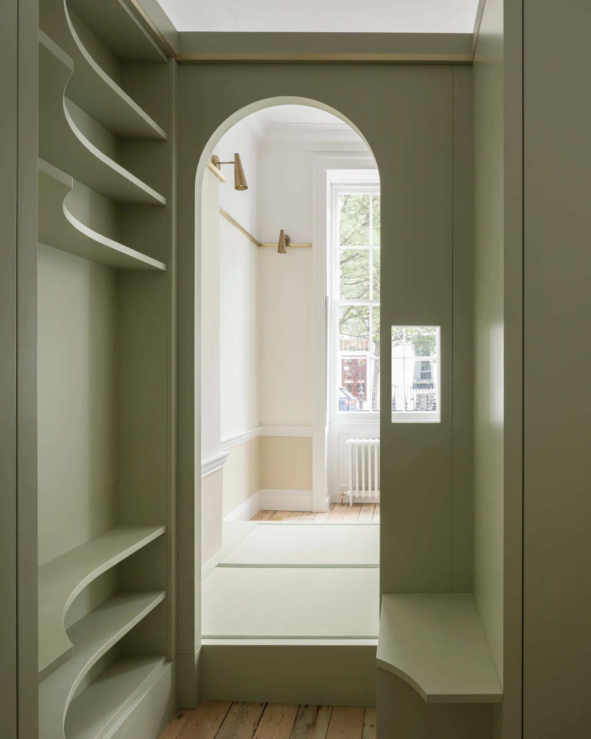 A sage green built-in storage unit with an arched entryway and curved shelves opens to a bright room with brass sconces and a sash window.
