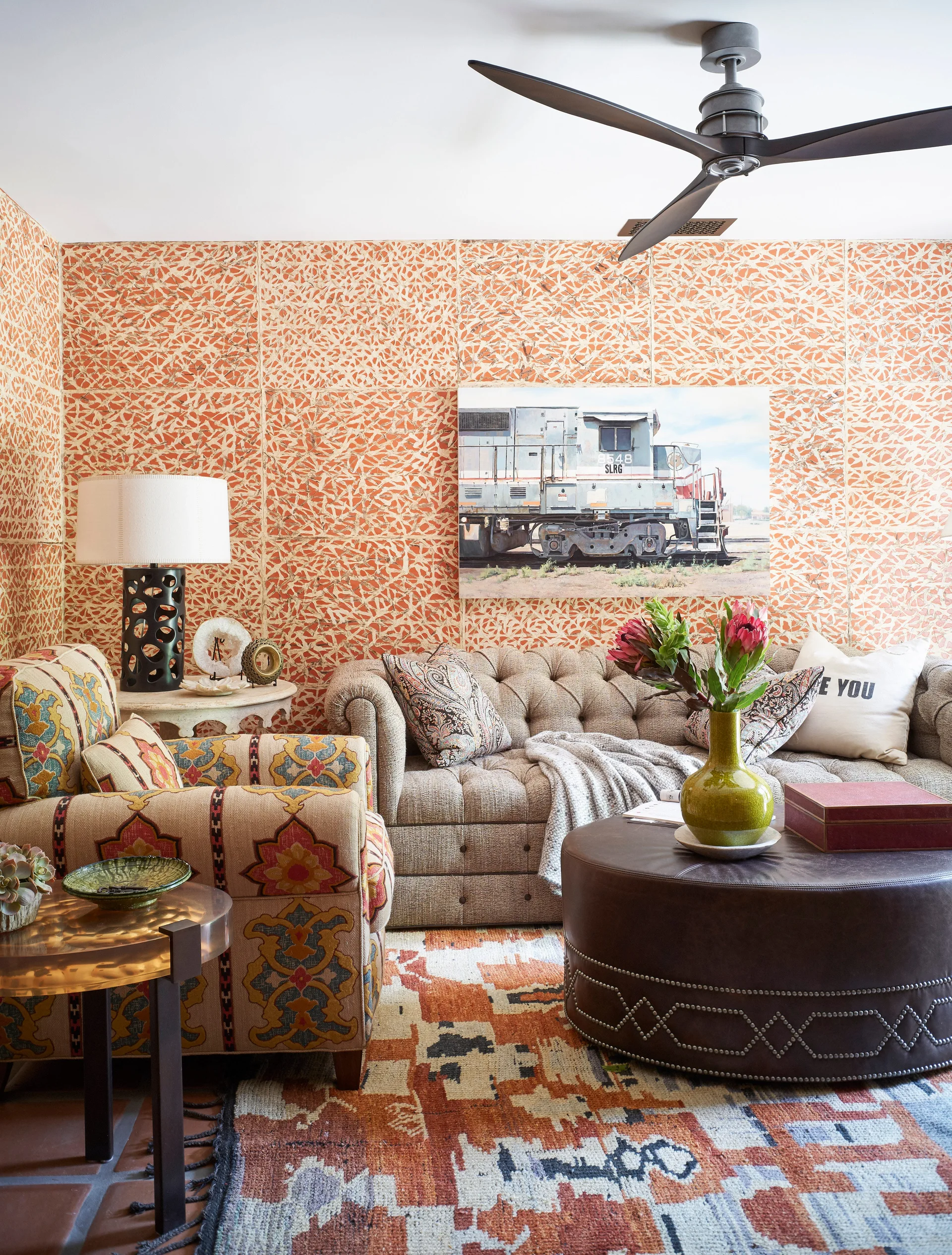 An eclectic living room features orange patterned wallpaper, a tufted neutral sofa, a patterned armchair, and a dark round leather ottoman.