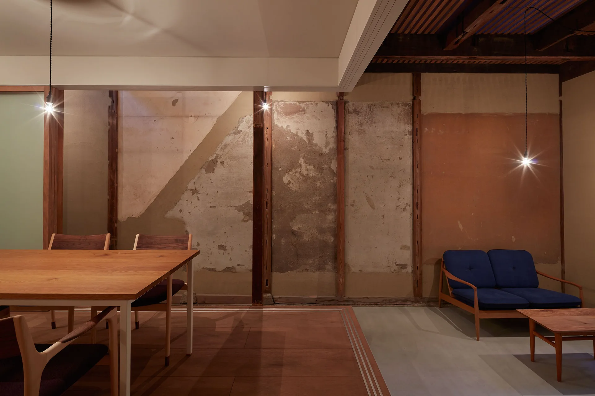 A wooden dining table sits next to a lounge area with a blue mid-century sofa set against a raw, unfinished plaster wall.