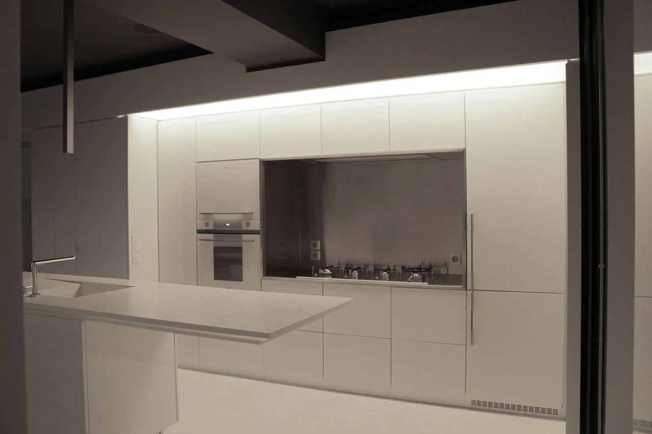 A minimalist white kitchen featuring handleless cabinetry, an integrated stainless steel stovetop alcove, and a matching white island under cove lighting.
