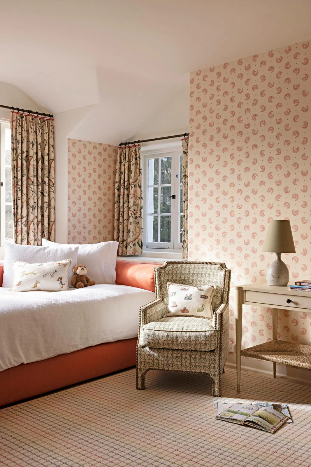 A cottage bedroom with patterned coral wallpaper, a green upholstered armchair, matching floral curtains, and a bed with white and coral bedding.