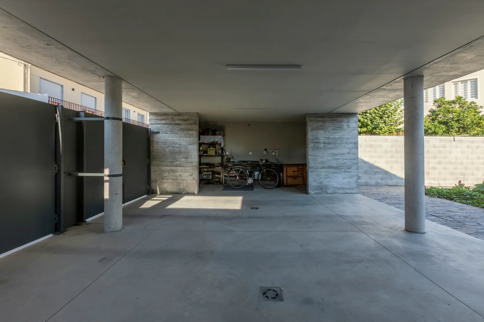 A minimalist concrete carport featuring round structural pillars, smooth gray flooring, and a rear utility alcove with a parked bicycle.