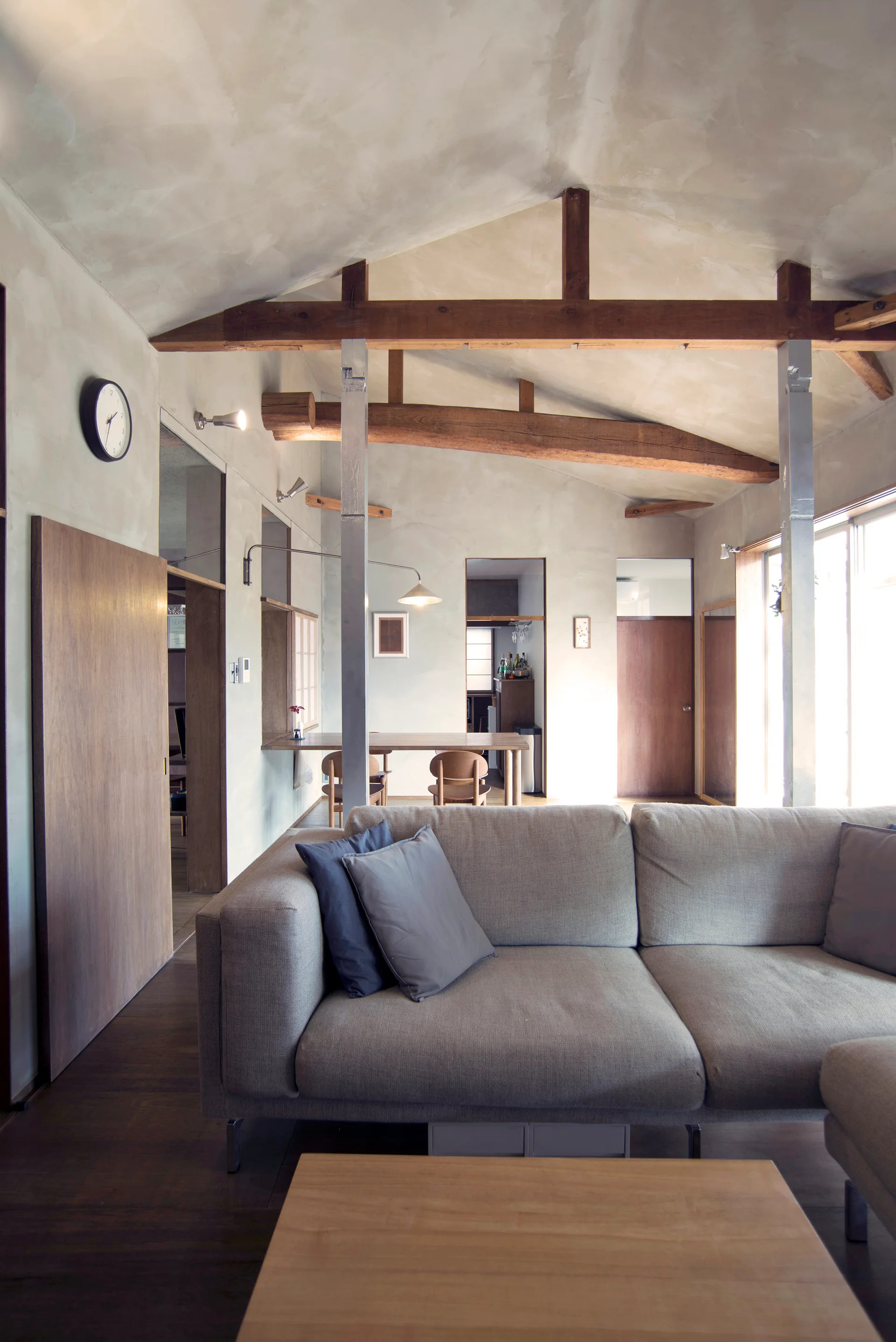 A grey sofa sits in a living room featuring textured plaster walls, exposed wooden ceiling beams, and a wooden sliding door.