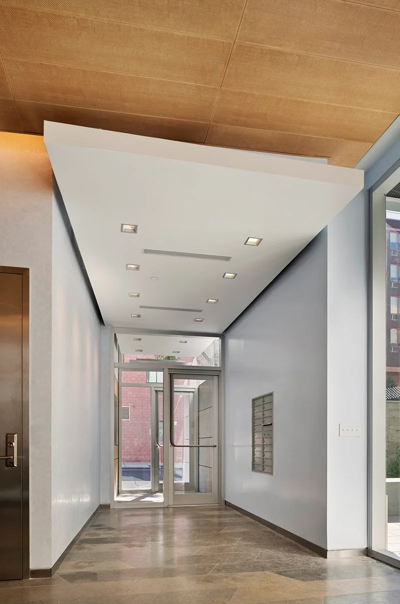 A modern building lobby featuring a white ceiling soffit with recessed lighting, stone-tiled floor, a wall-mounted mailbox, and glass entry doors.