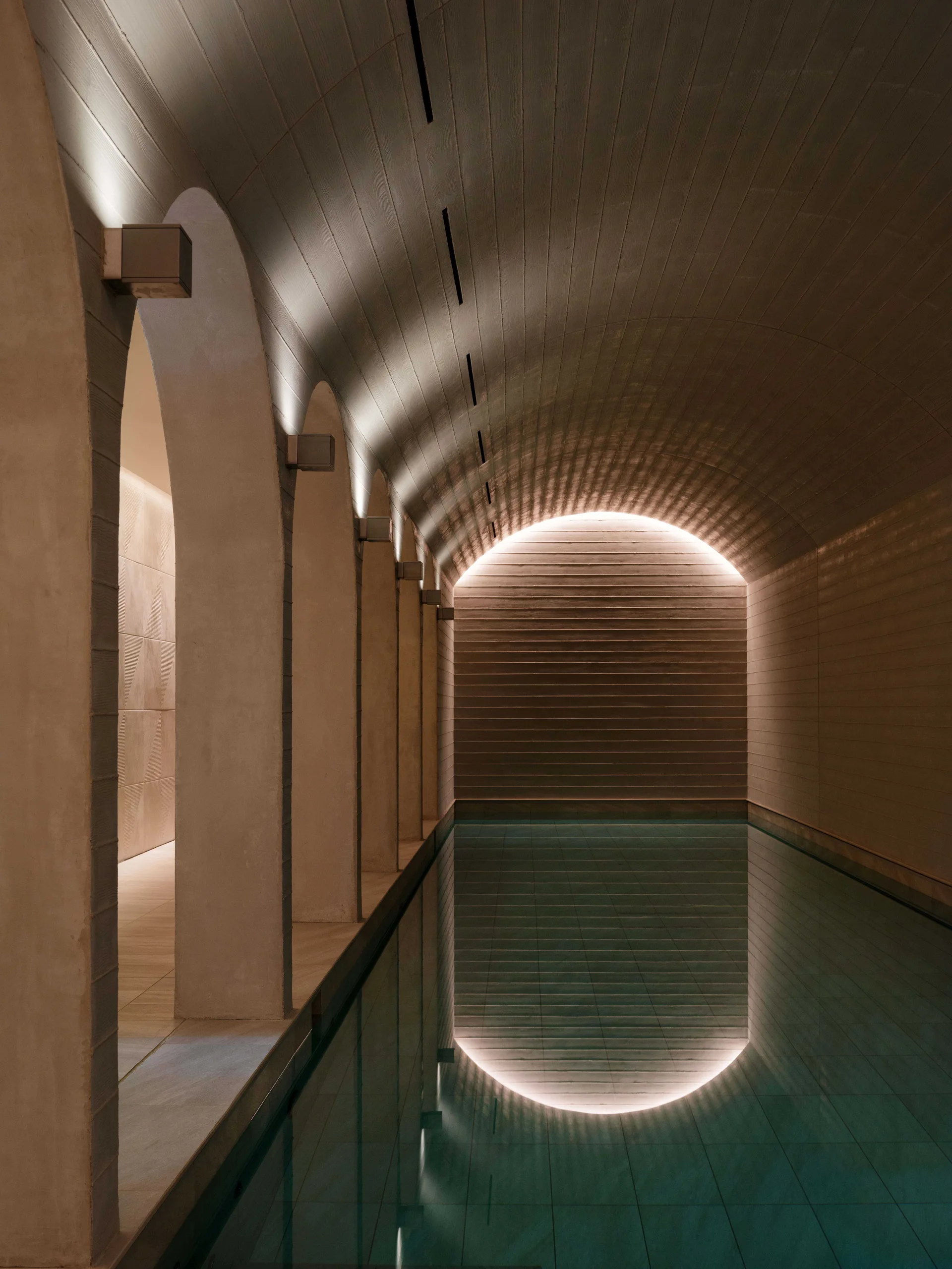 An indoor swimming pool sits beneath a curved, vaulted concrete ceiling supported by a row of stone-like arches with soft architectural lighting.