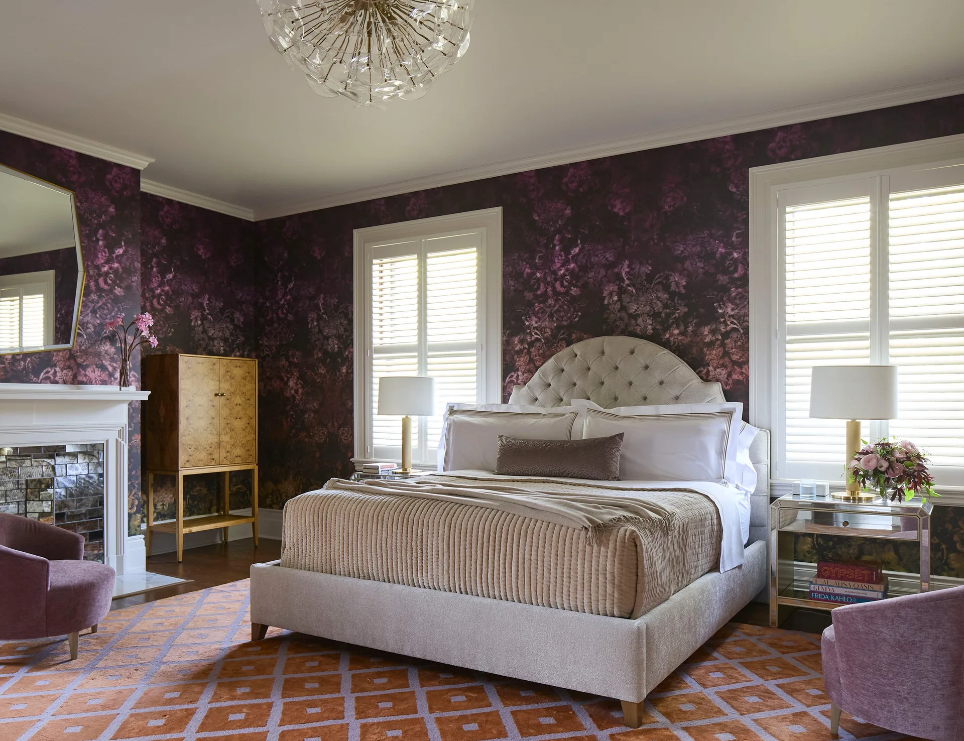 A residential bedroom featuring deep plum floral wallpaper, a beige tufted headboard, white plantation shutters, and a geometric orange and purple rug.
