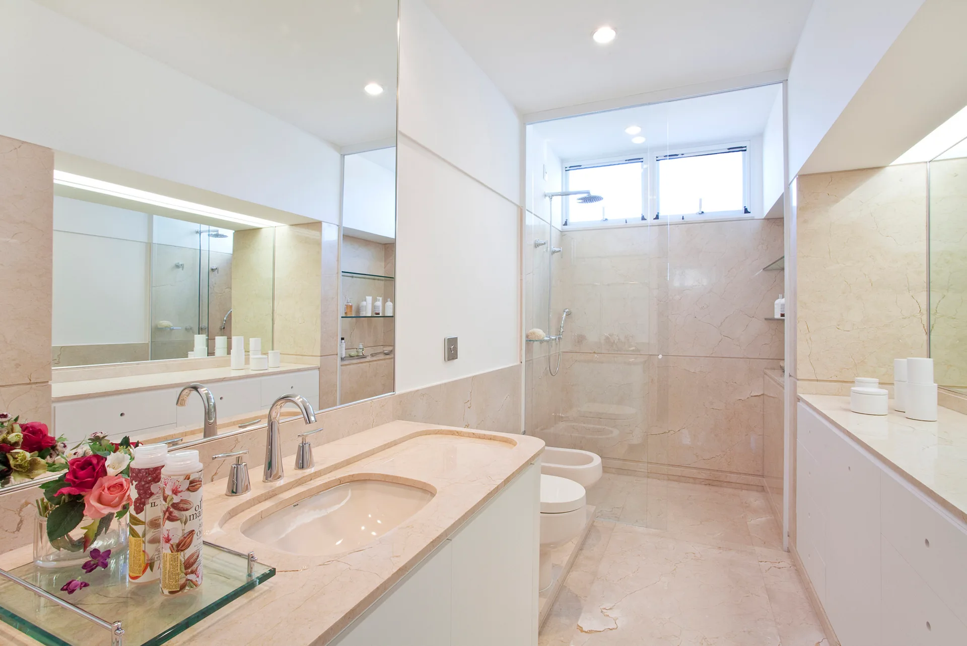 A modern bathroom features beige veined stone walls, a matching marble vanity with chrome faucets, and a glass-enclosed walk-in shower.