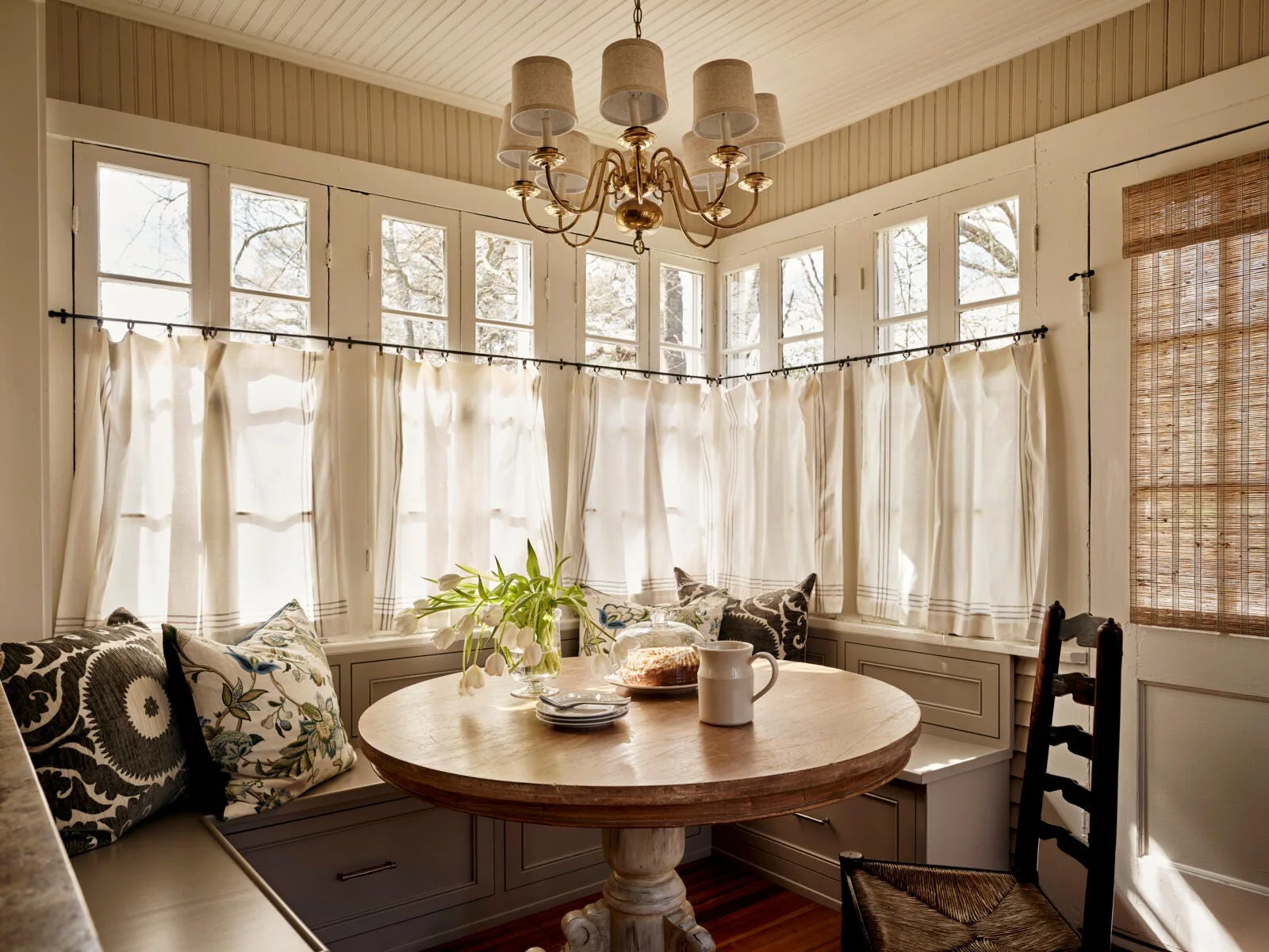 A cozy breakfast nook features a round wooden pedestal table, a grey built-in corner banquette with patterned pillows, and white cafe curtains.