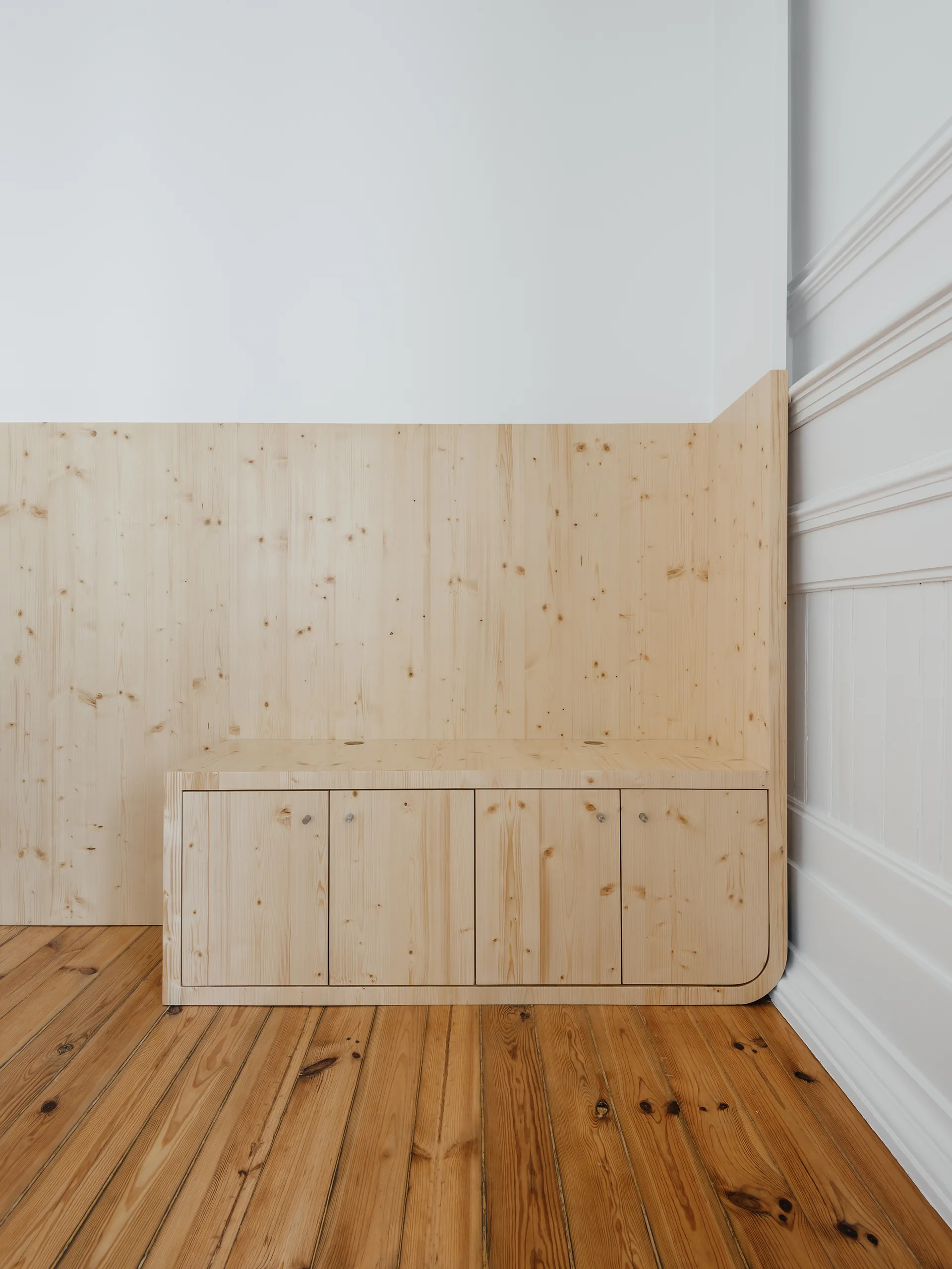 A low built-in pine cabinet with integrated wall paneling sits on a wood plank floor next to a white wall with decorative trim.