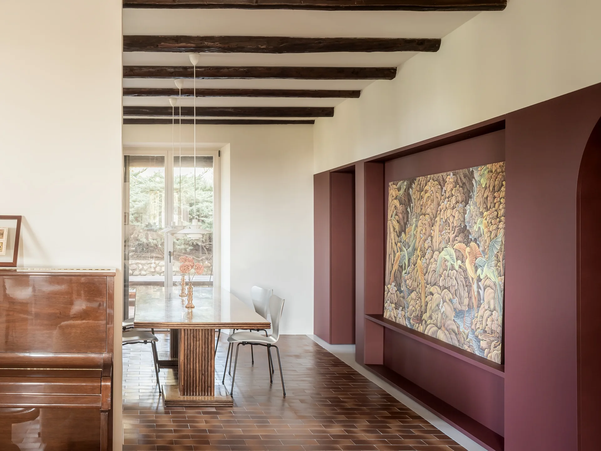 A dining room features a wooden table, dark ceiling beams, a brown tiled floor, and a large burgundy wall niche showcasing a landscape artwork.