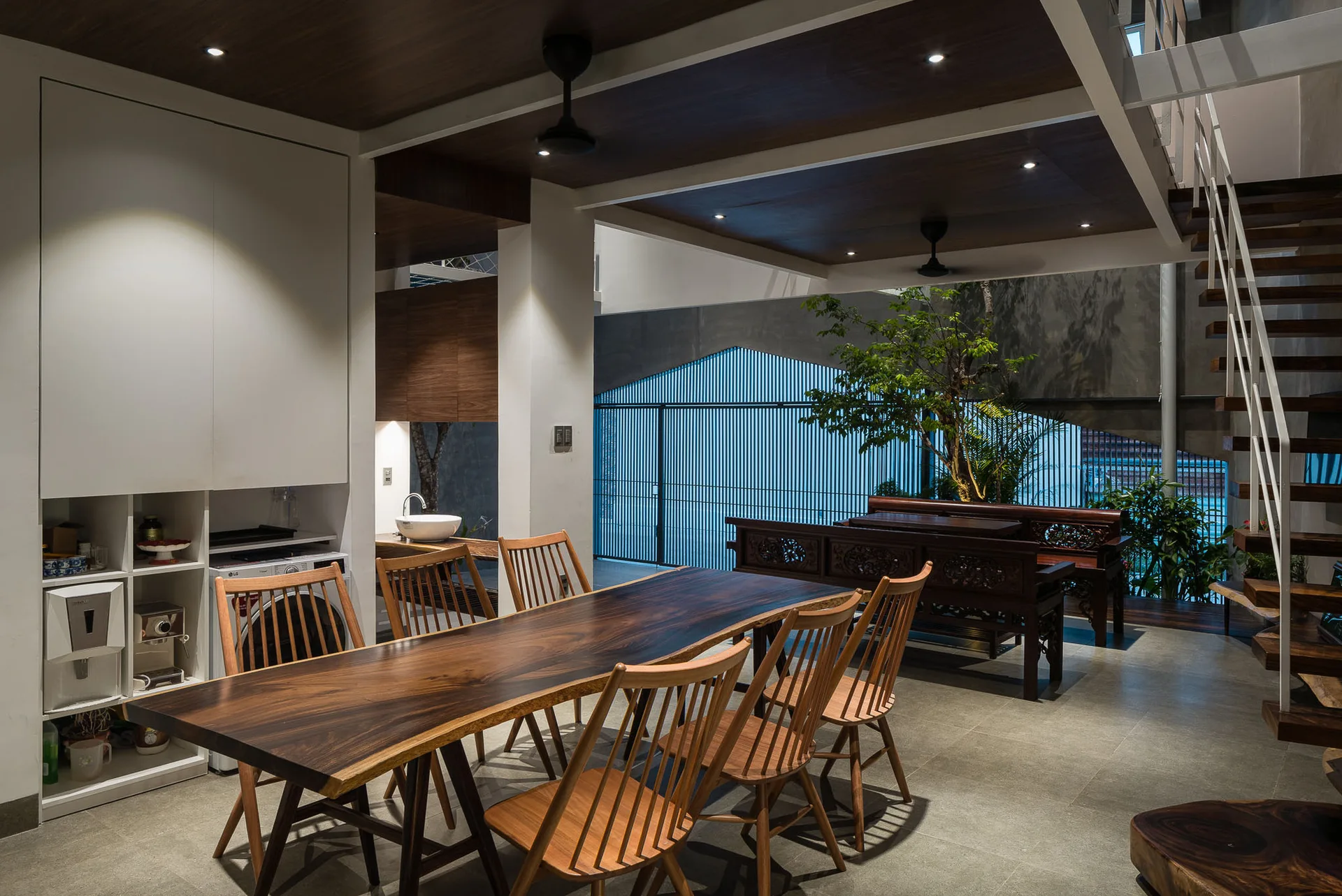 A contemporary open-plan dining room features a long live-edge wood table surrounded by wooden spindle-back chairs under a paneled ceiling.