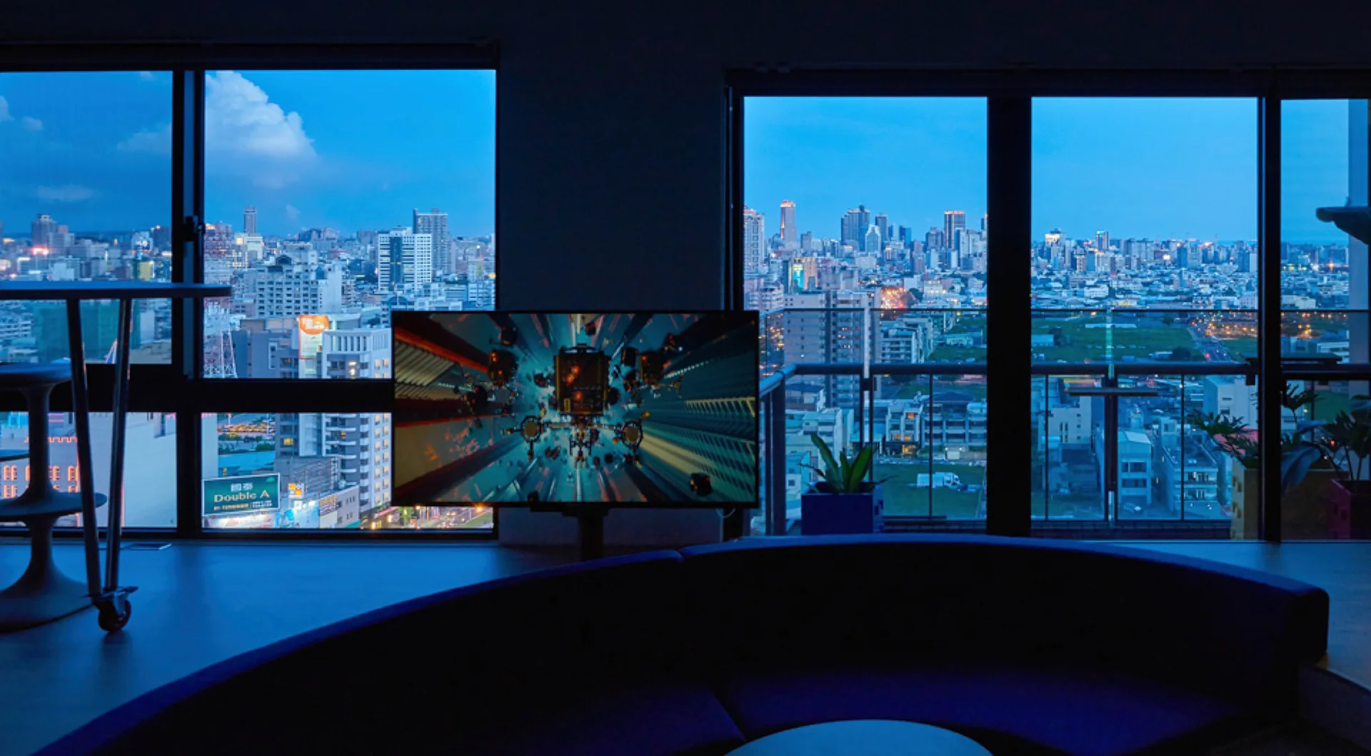 A dark modern living room features a curved blue sofa facing a television screen against large windows with a twilight city view.