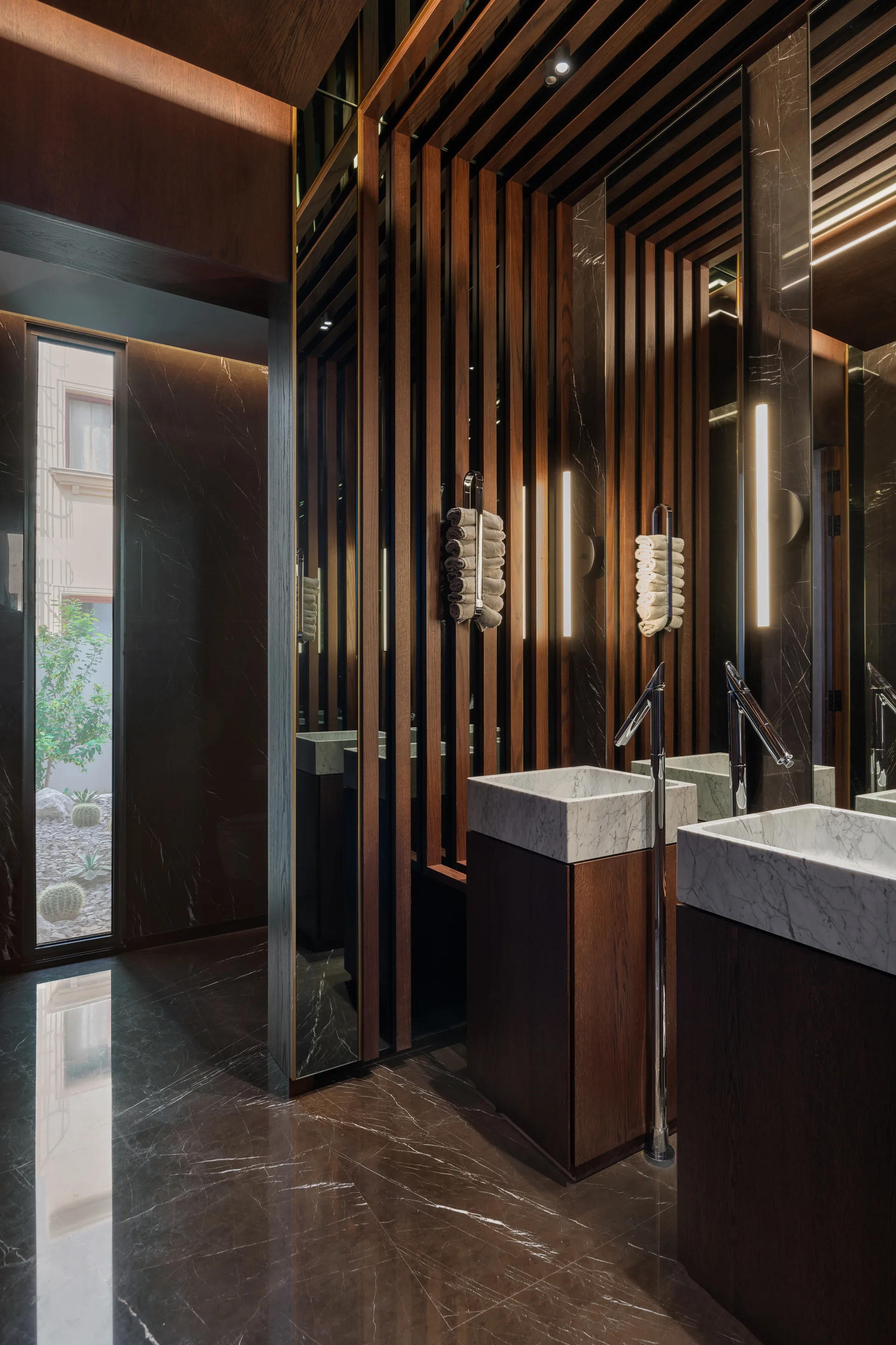 A modern bathroom features dark wood vertical slats, two block marble vanities with floor-mounted faucets, and reflective mirror-paneled walls.