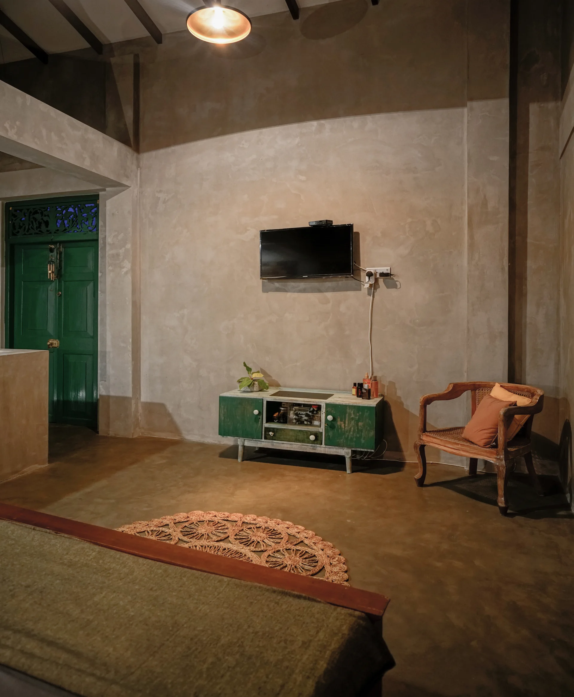 A rustic bedroom features textured concrete walls, a green distressed media console under a wall-mounted TV, a wooden armchair, and a circular jute rug.