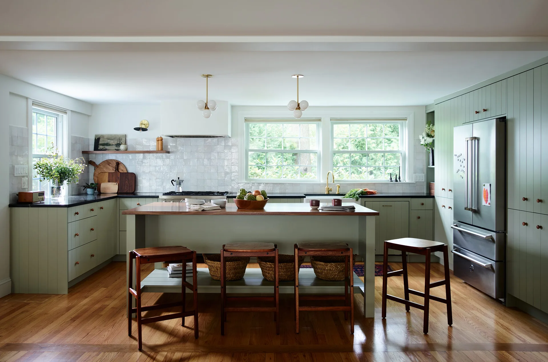 A residential kitchen features sage green cabinetry, a central wood-topped island with wooden stools, and a glossy light gray tiled backsplash.