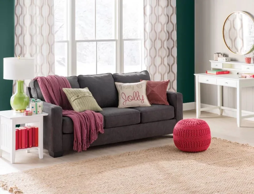 A charcoal grey sofa with colorful pink and green accent pillows sits on a jute rug next to a white side table and a pink knitted pouf.