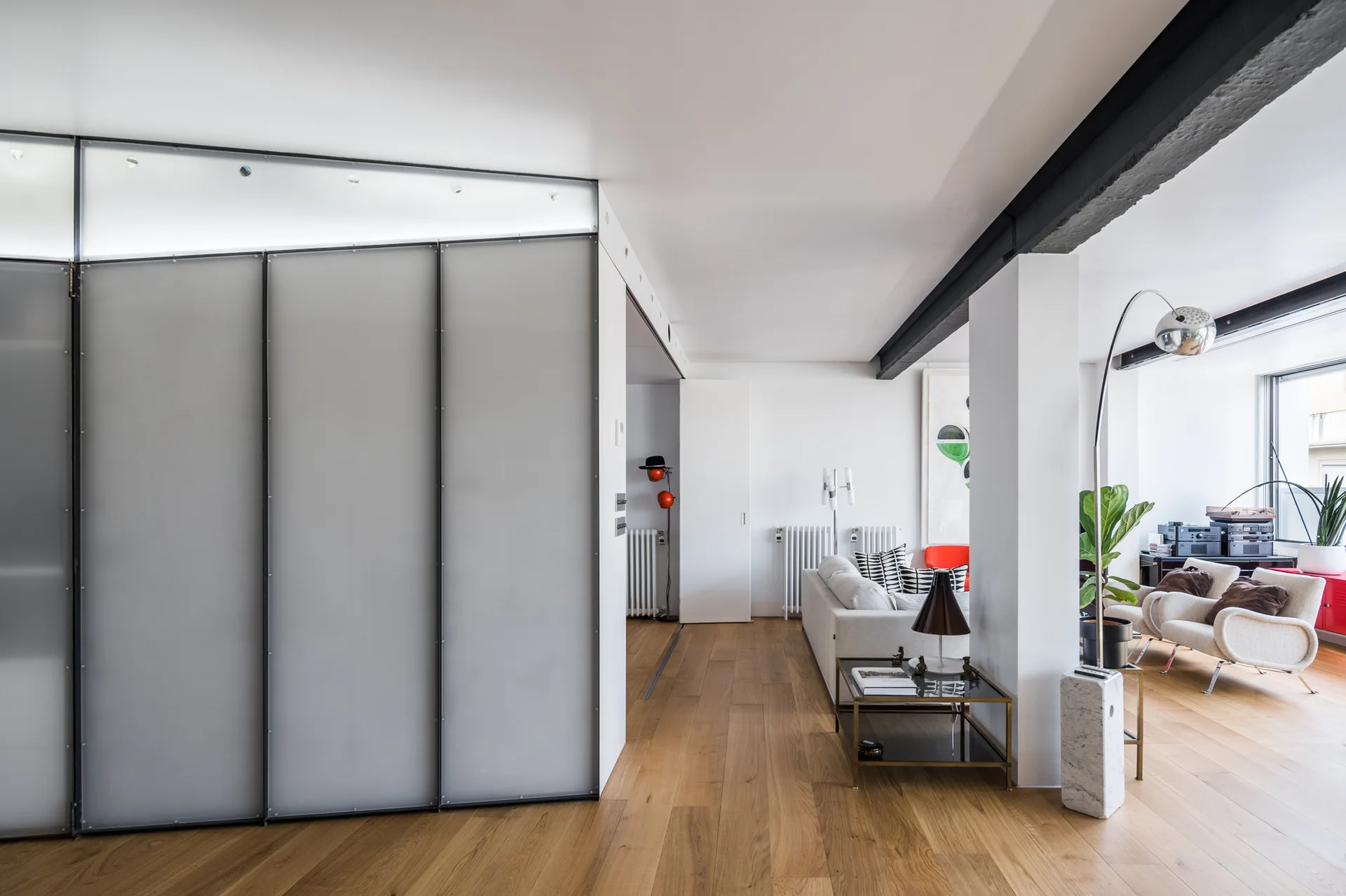 A minimalist living room features wood flooring, a large frosted glass sliding partition, a white sofa, and a black ceiling beam.