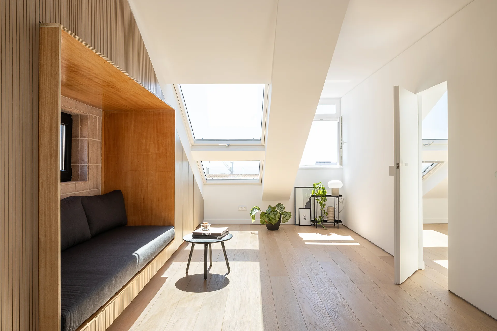 A minimalist attic living room featuring a built-in wooden alcove bench with a dark cushion, light wood floors, and two bright skylights.
