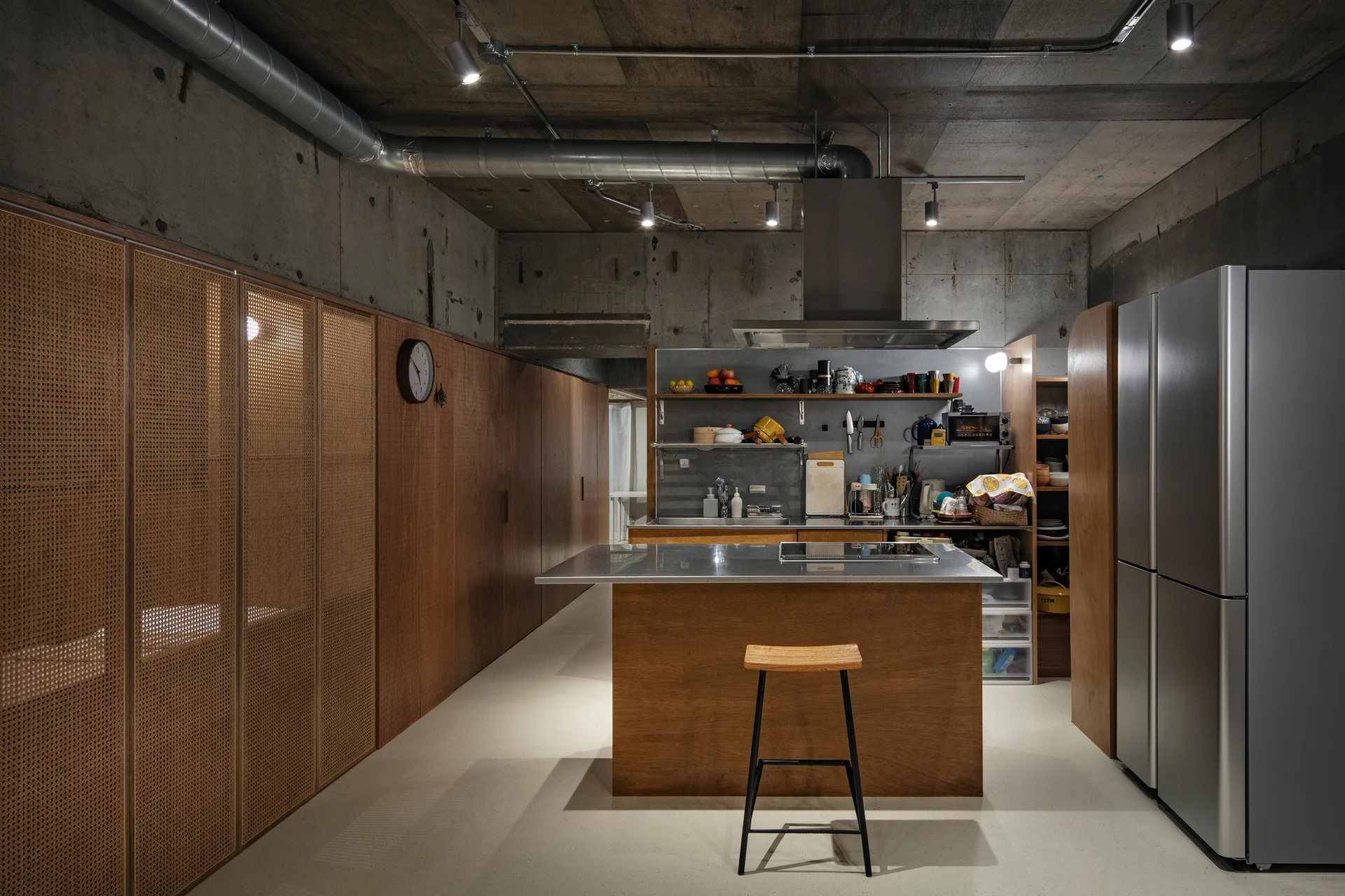 An industrial kitchen features exposed concrete walls, a wooden island with a stainless steel top, and a wall of cabinets with woven cane panels.