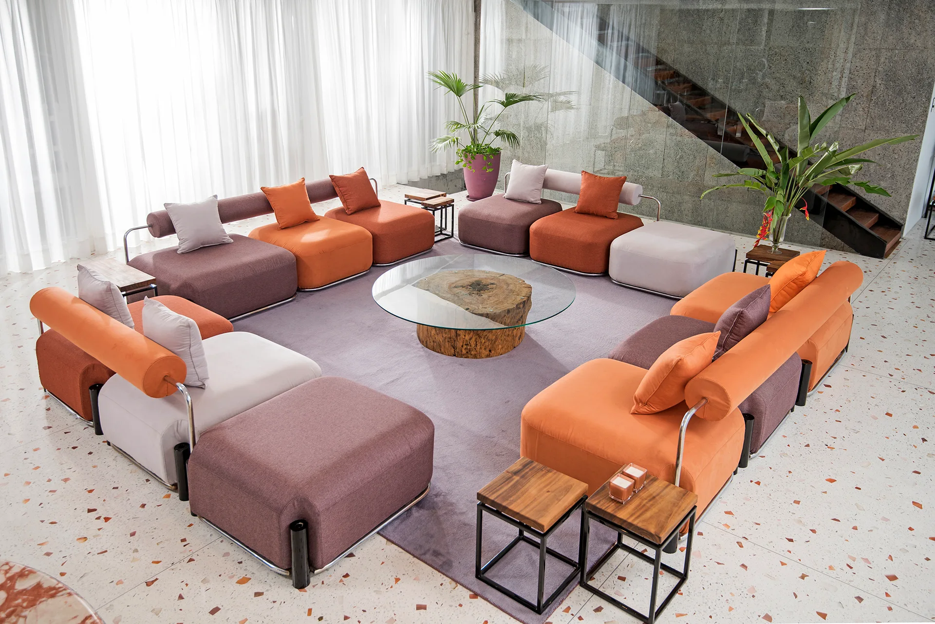A large circular modular sofa in terracotta and lavender tones surrounds a glass-topped log coffee table on white terrazzo flooring.