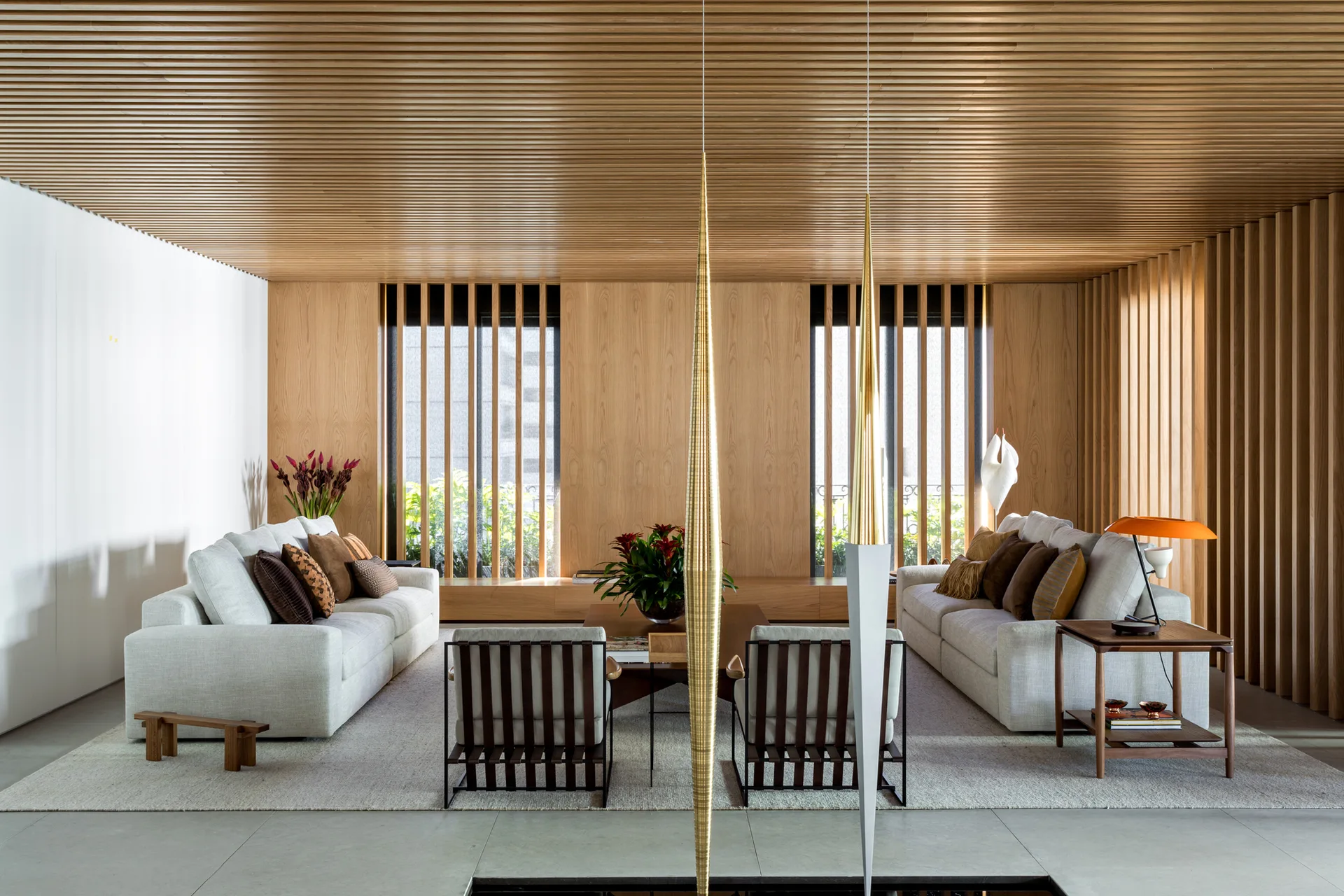 A spacious living room with two beige sofas, two wooden armchairs, a large area rug, and a continuous wood-slatted ceiling.