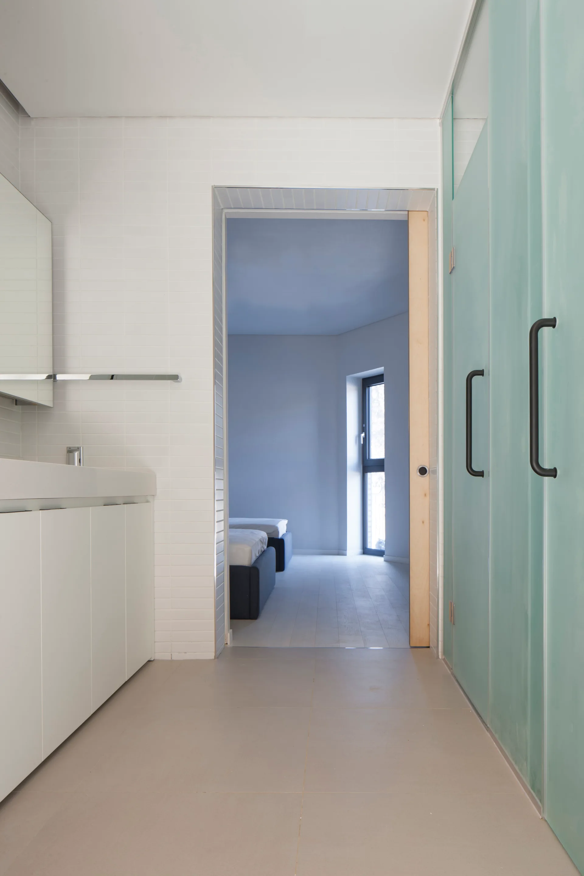 A minimalist bathroom with a white vanity and teal frosted glass cabinets leads through a wood-trimmed doorway into an adjacent bedroom.