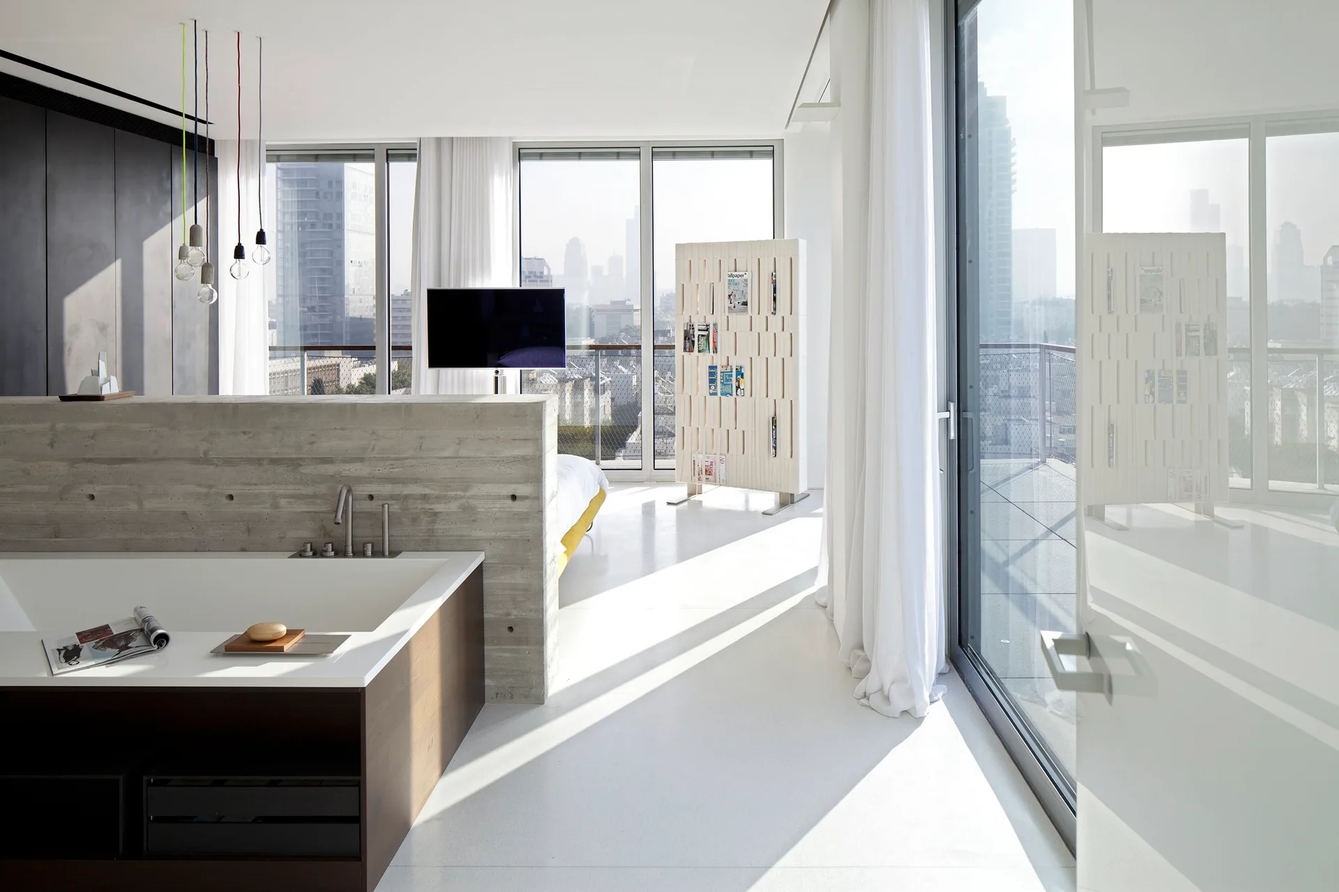 An open-plan bedroom featuring an integrated wood-framed bathtub separated from the sleeping area by a low, raw concrete partition wall.
