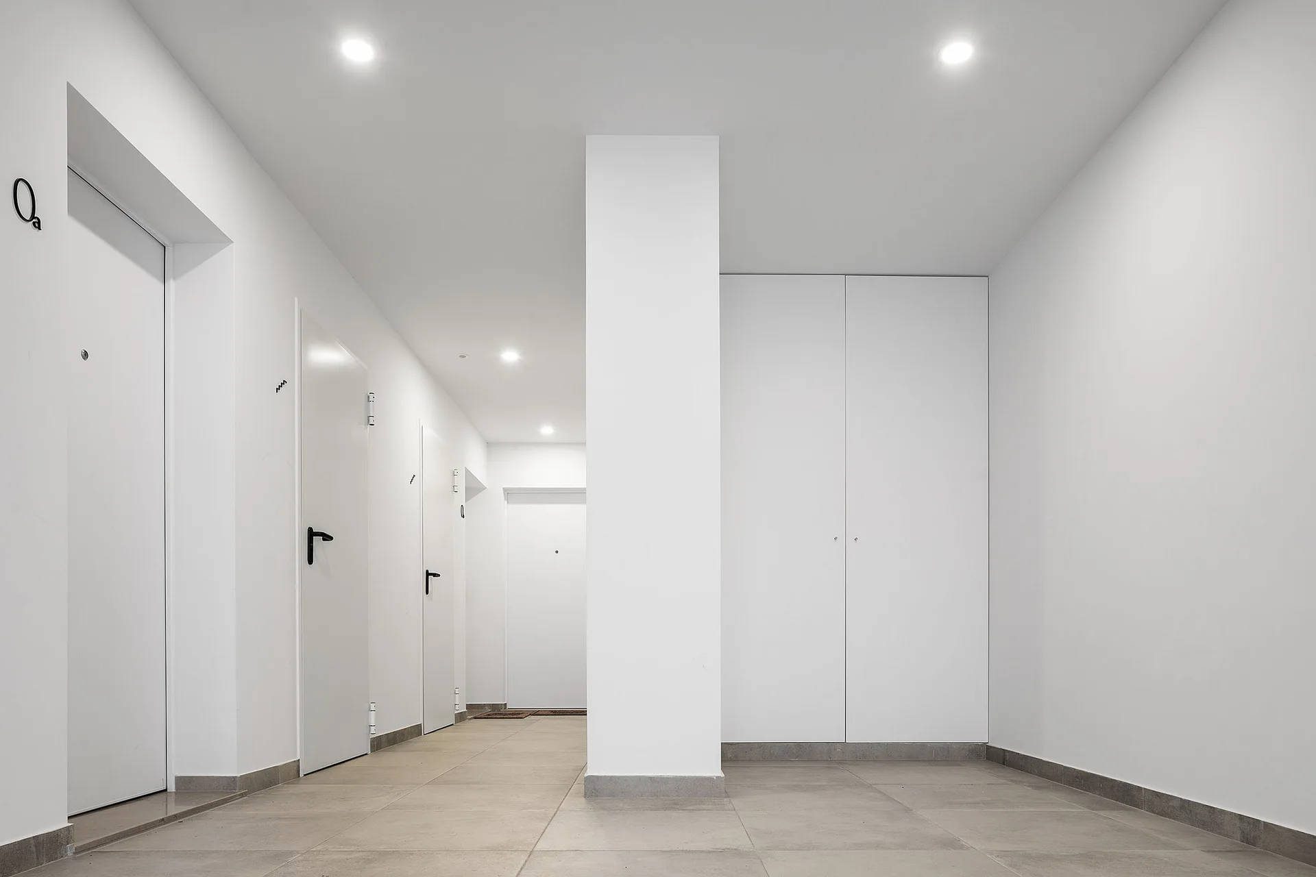 A minimalist white hallway featuring flush doors with black handles, a central structural pillar, and light grey tile flooring.