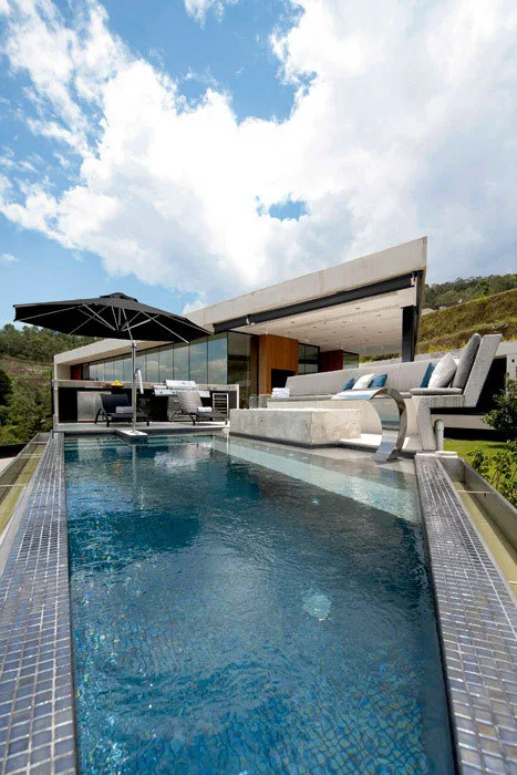 A modern concrete patio features a rectangular swimming pool, a long built-in sofa, a stainless steel grill, and a black cantilever umbrella.