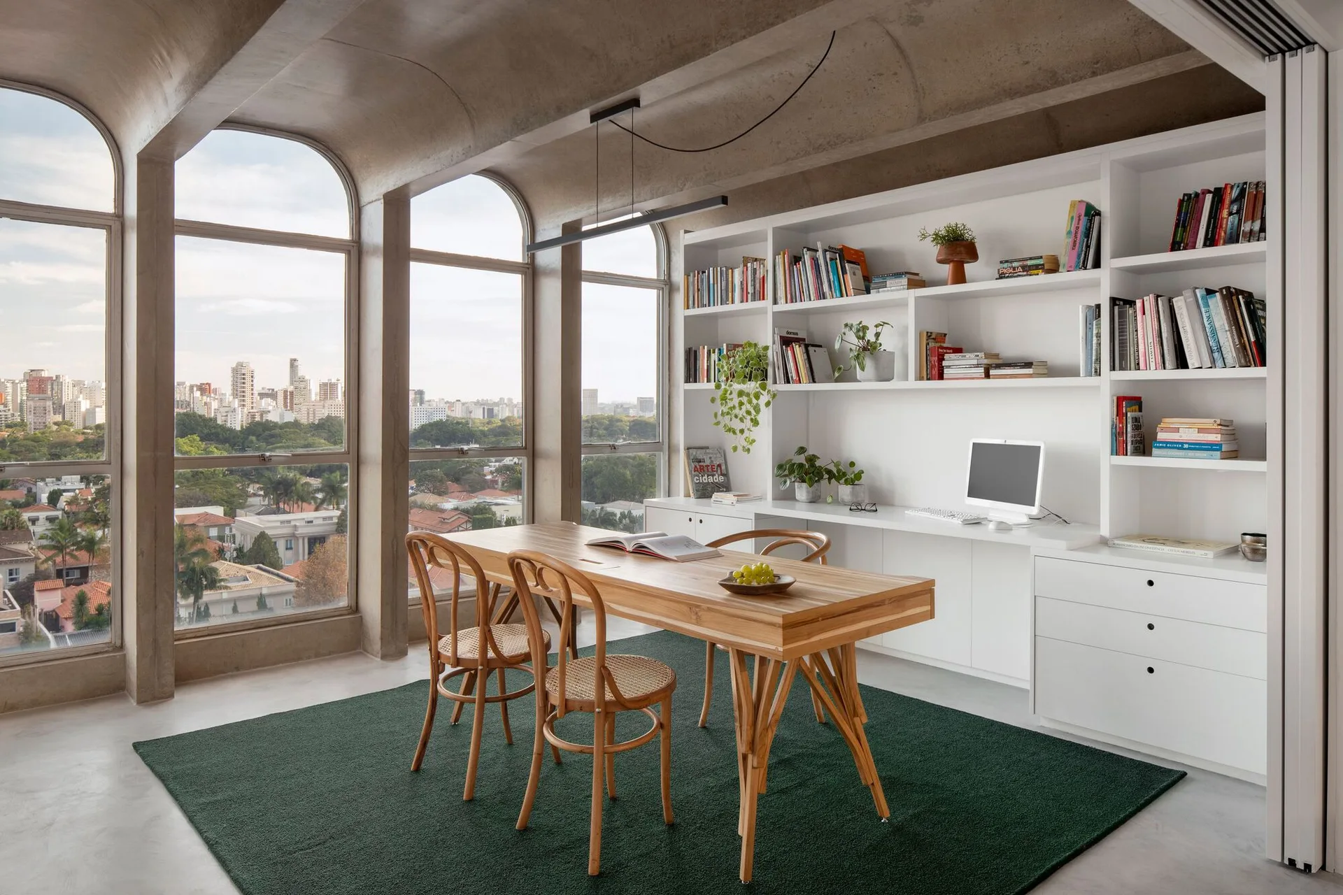 A modern room featuring a wooden table on a green rug beneath a vaulted concrete ceiling, with white built-in shelving and arched windows.