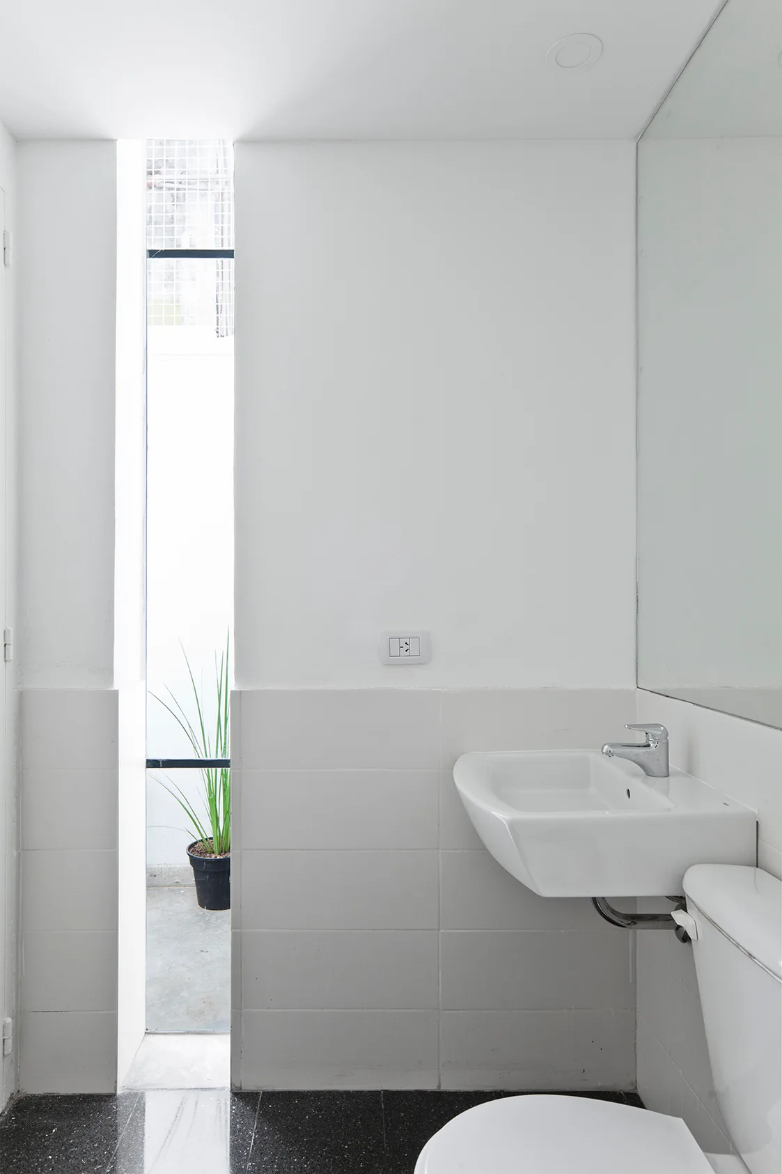 A minimalist bathroom features white tiled walls, a wall-mounted sink, dark speckled flooring, and a narrow vertical window revealing a plant outside.