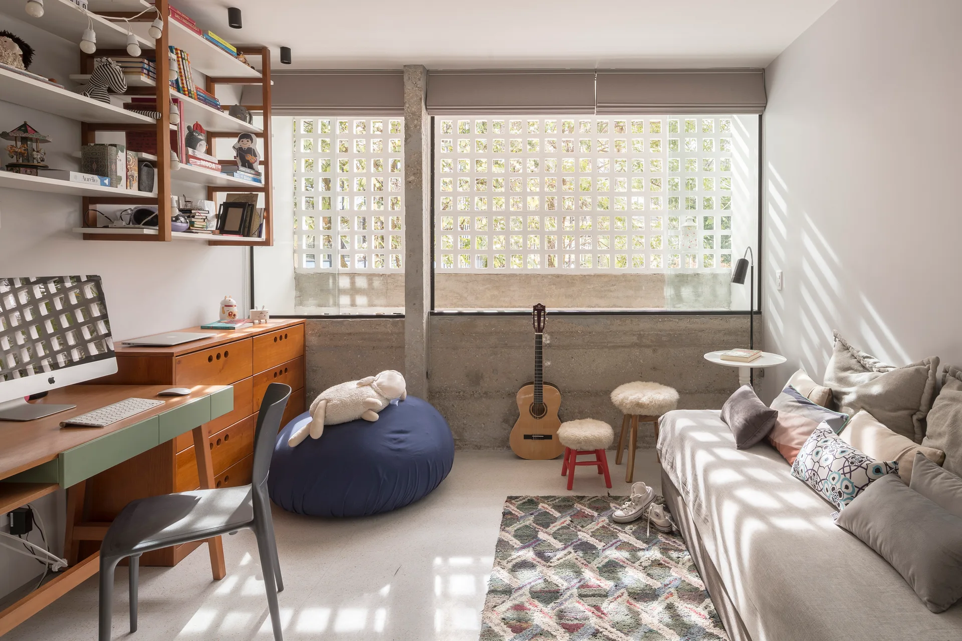 A youth bedroom with a wooden desk and chair, a grey sofa, and a large window with a concrete lattice screen casting gridded shadows.