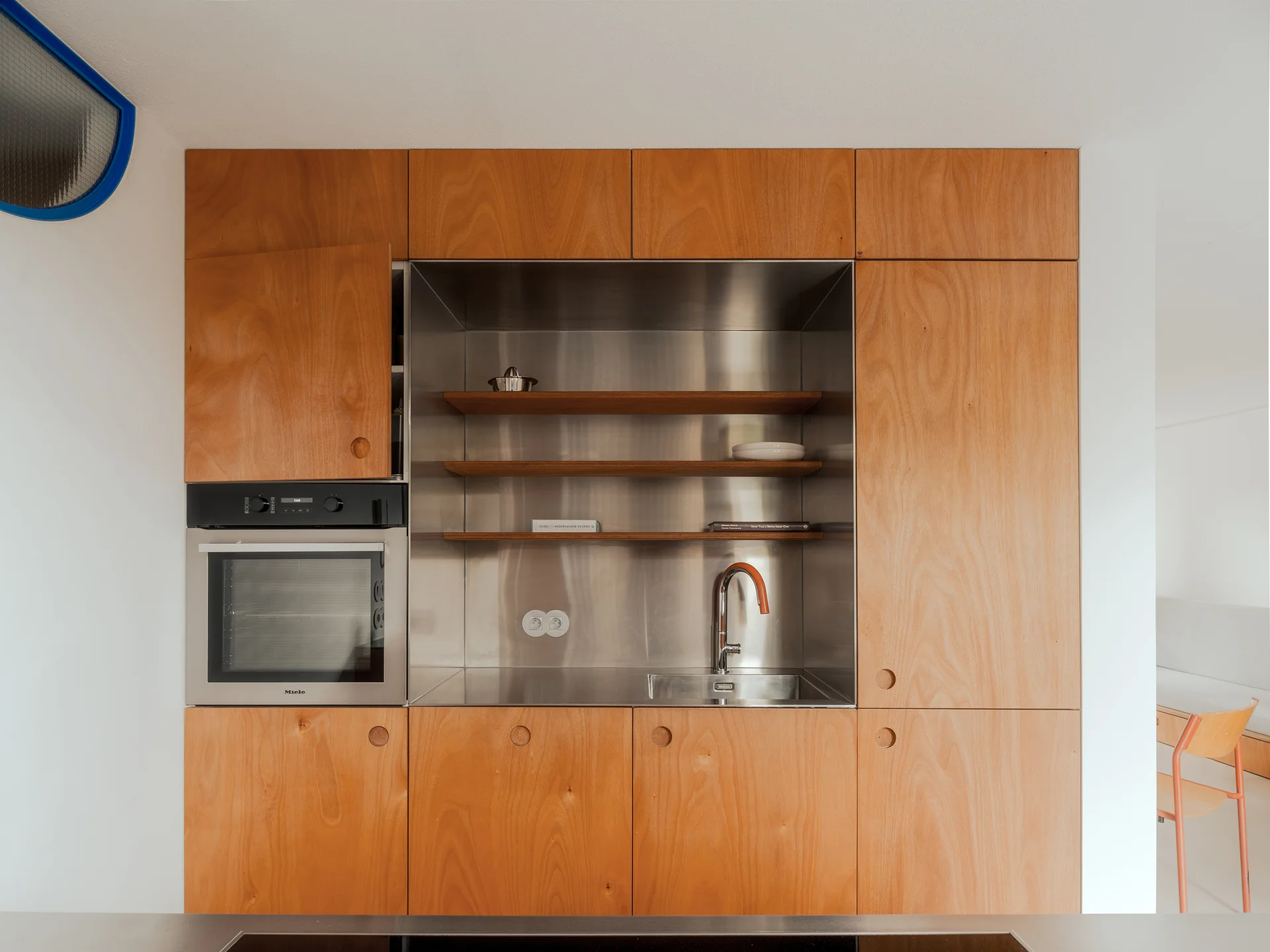 A flat-front plywood kitchen cabinet wall features a recessed stainless steel sink workstation, built-in wall oven, and minimalist open shelving.