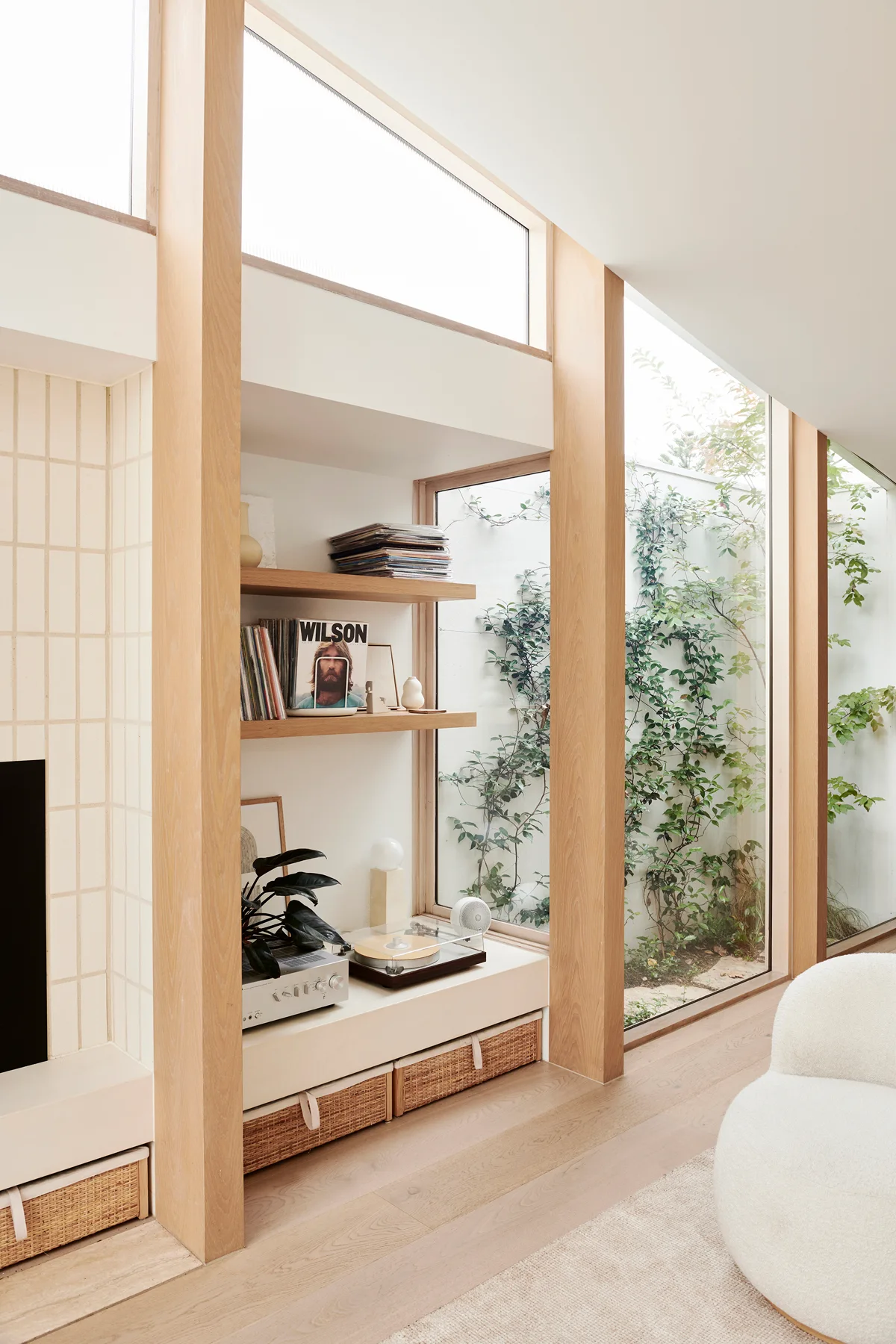 Light wood built-in shelving holding records and a turntable sits next to a large glass window overlooking a green courtyard garden.