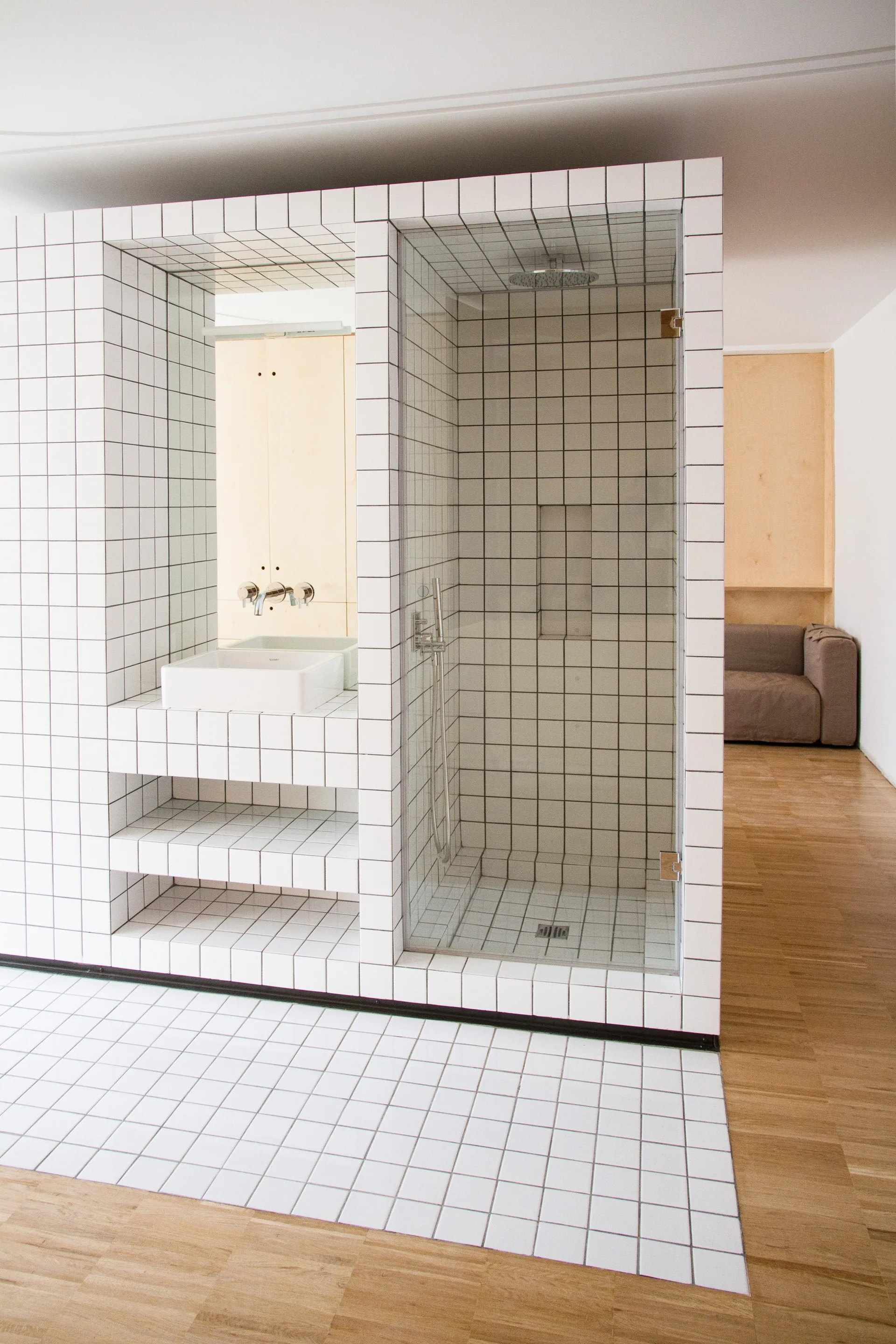 A central white square-tiled structure contains a bathroom vanity and a walk-in shower next to light wood flooring in an open space.
