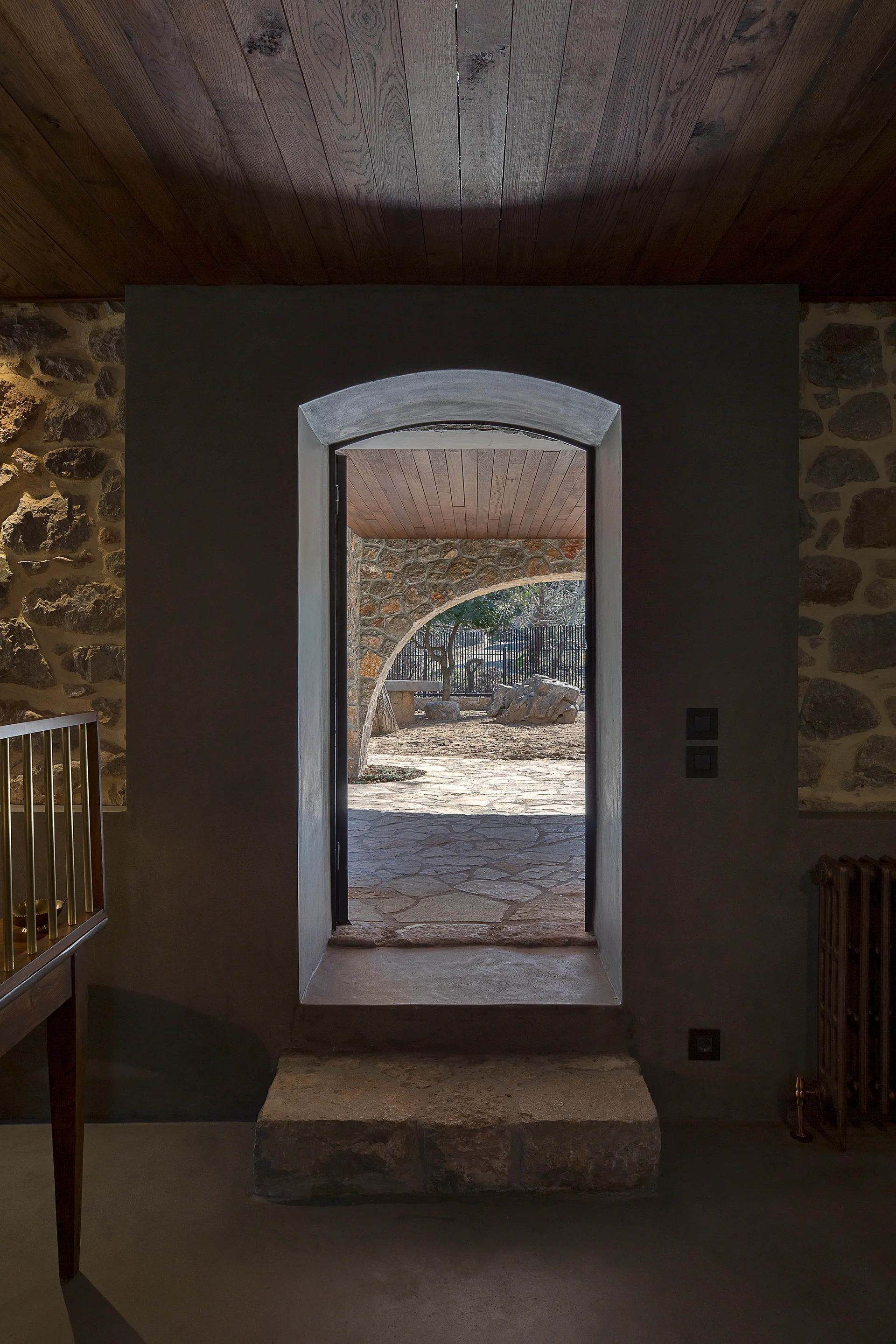An arched plaster doorway with a rustic stone step leads to an outdoor courtyard beneath a dark wood plank ceiling.