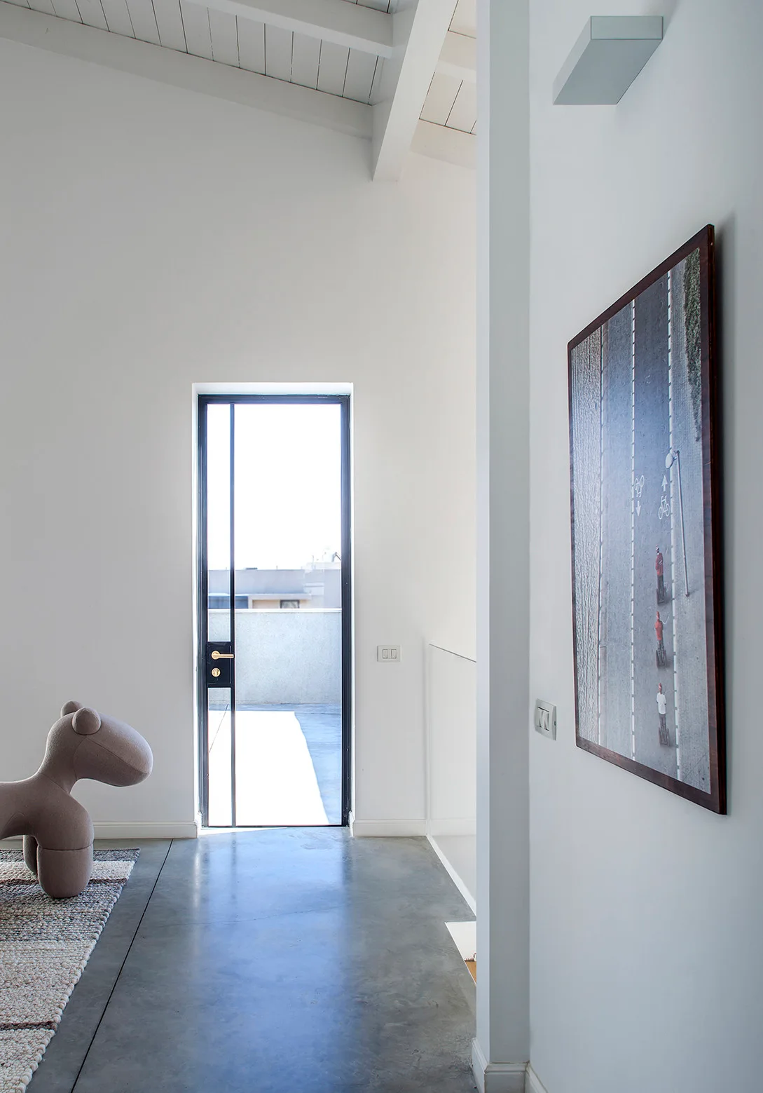 A minimalist white hallway featuring a black-framed glass balcony door, matte concrete floors, and a vertical framed print.