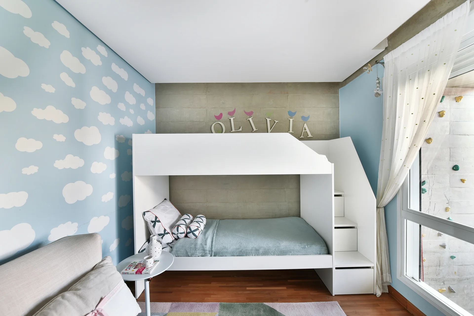 A modern children's bedroom features a white wood bunk bed set against a concrete accent wall and light blue cloud wallpaper.
