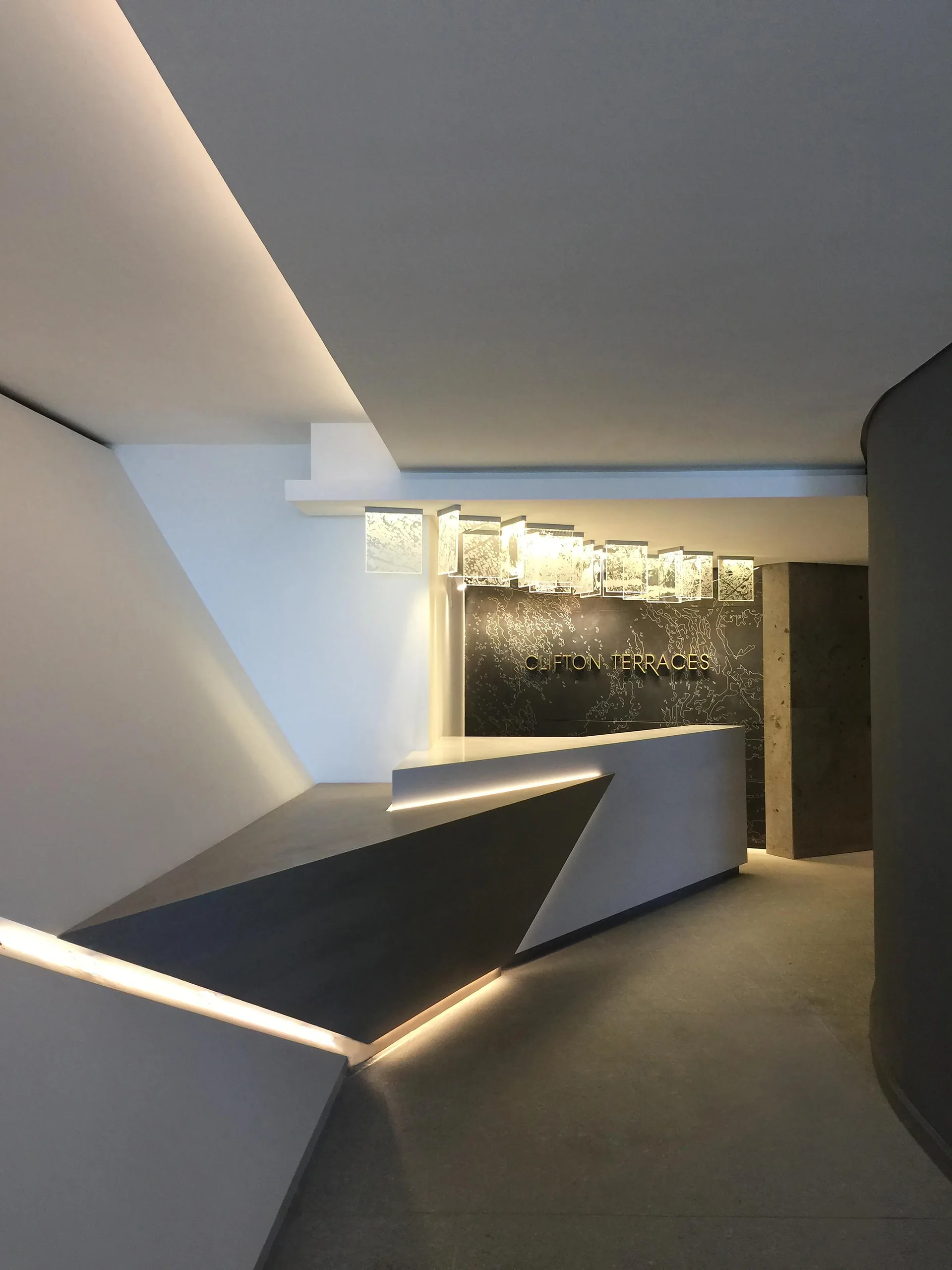 A modern reception lobby featuring a white geometric desk with integrated LED lighting, a dark patterned accent wall, and hanging glass pendant lights.