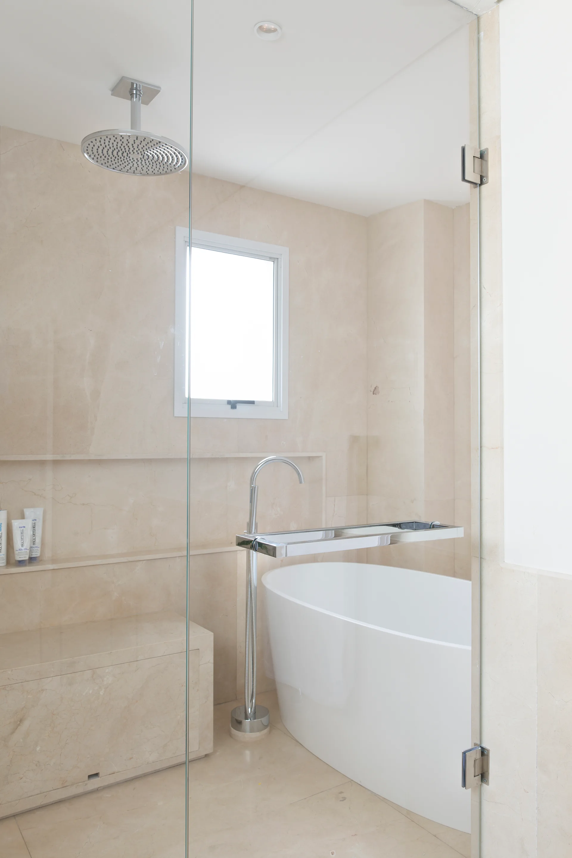 A modern bathroom with beige marble tile walls features a white freestanding bathtub next to a glass-enclosed shower with a rain shower head.