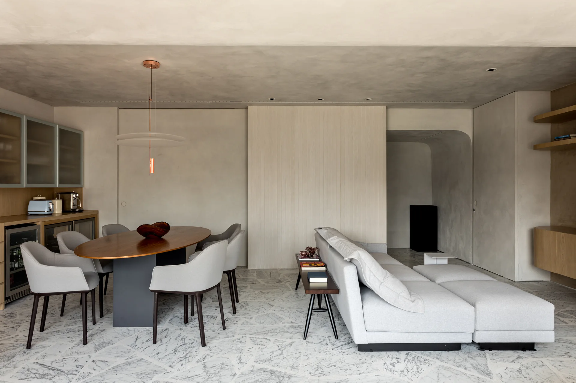 An open-plan living and dining area featuring a modular grey sofa, an oval wood table, grey chairs, and a distinctive grey and white stone tile floor.