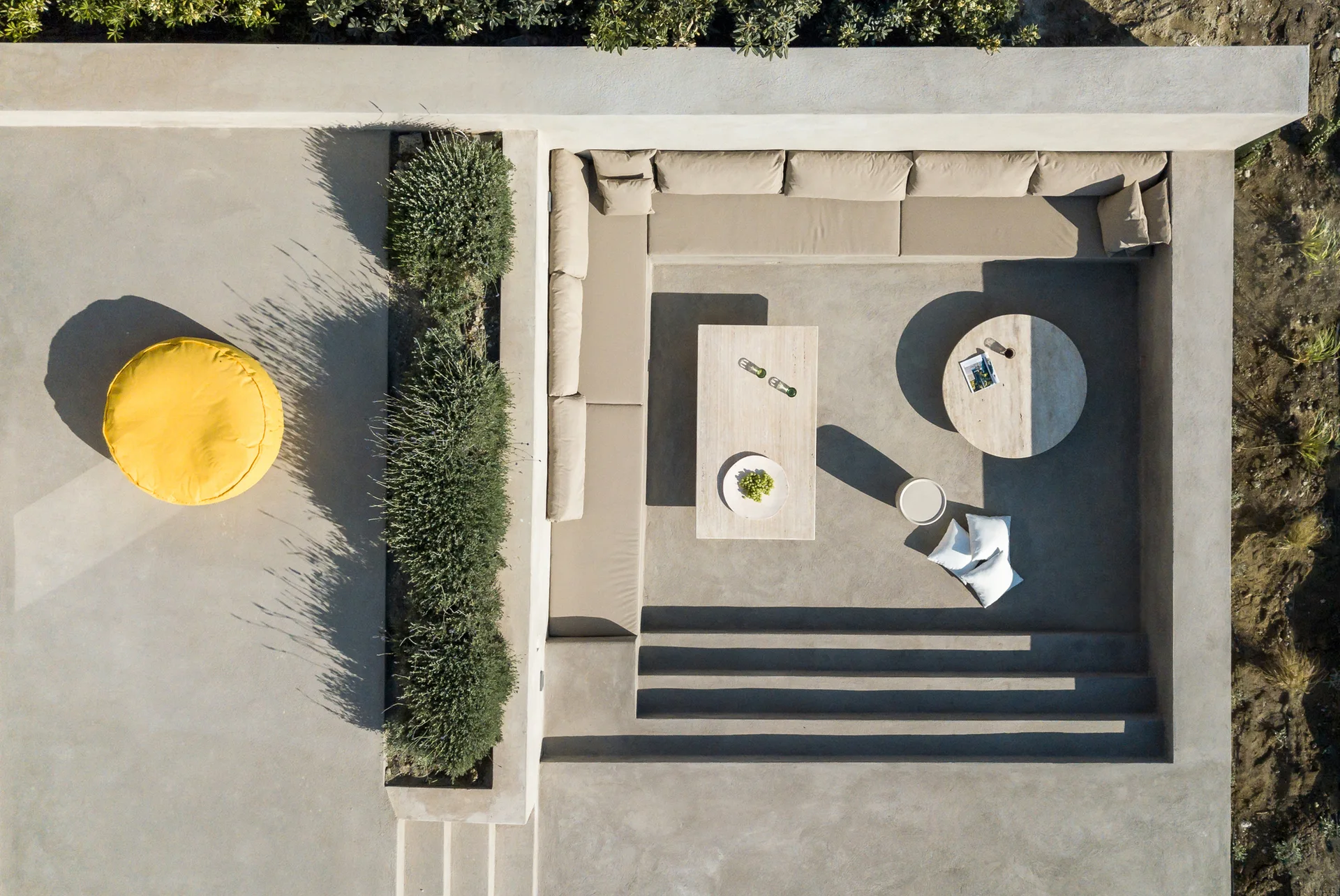 An aerial view of a sunken concrete patio featuring L-shaped built-in bench seating with beige cushions, stone tables, and a bright yellow floor cushion.