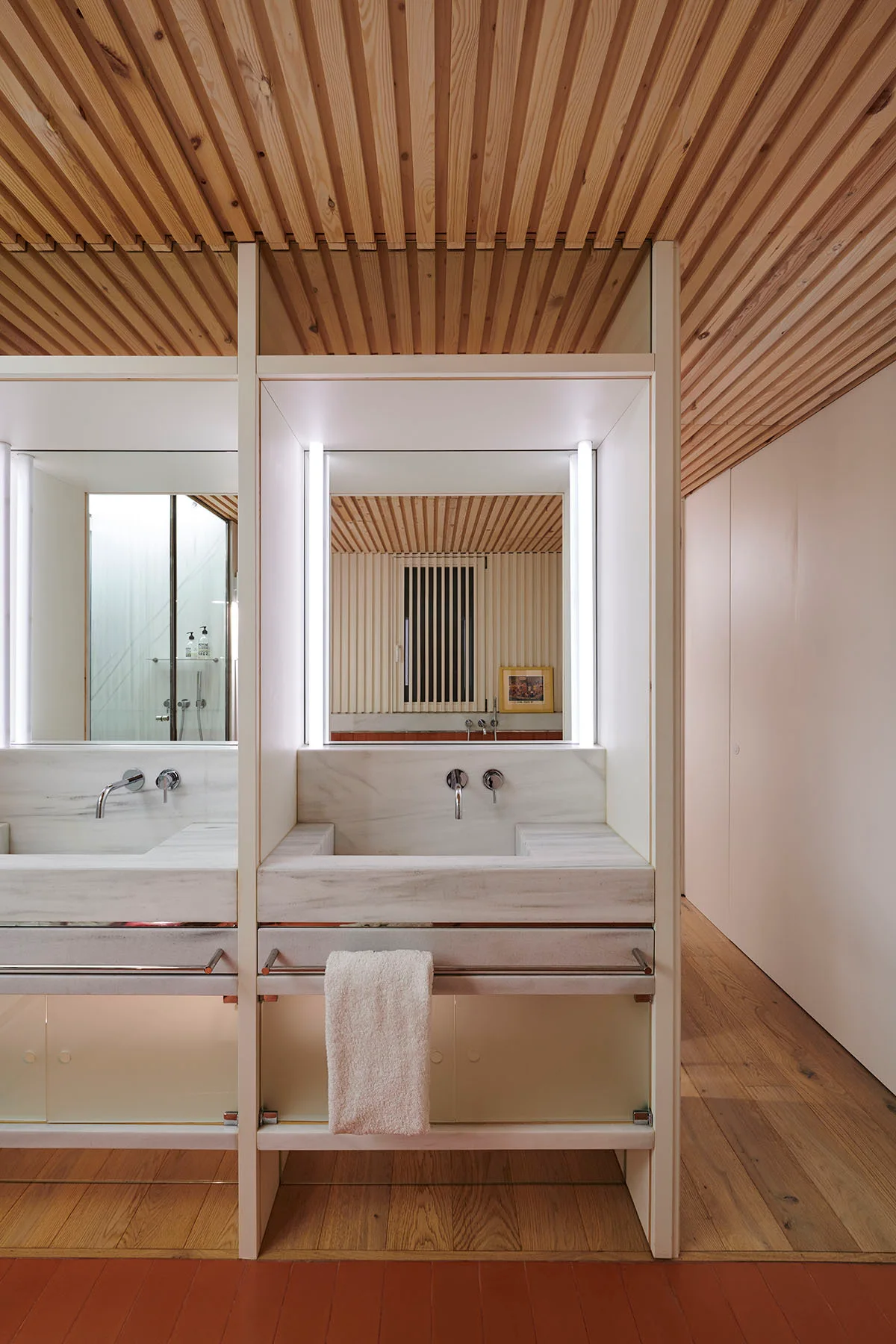 A modern bathroom features a double marble vanity with wall-mounted faucets beneath a linear wood slatted ceiling and integrated mirror lighting.