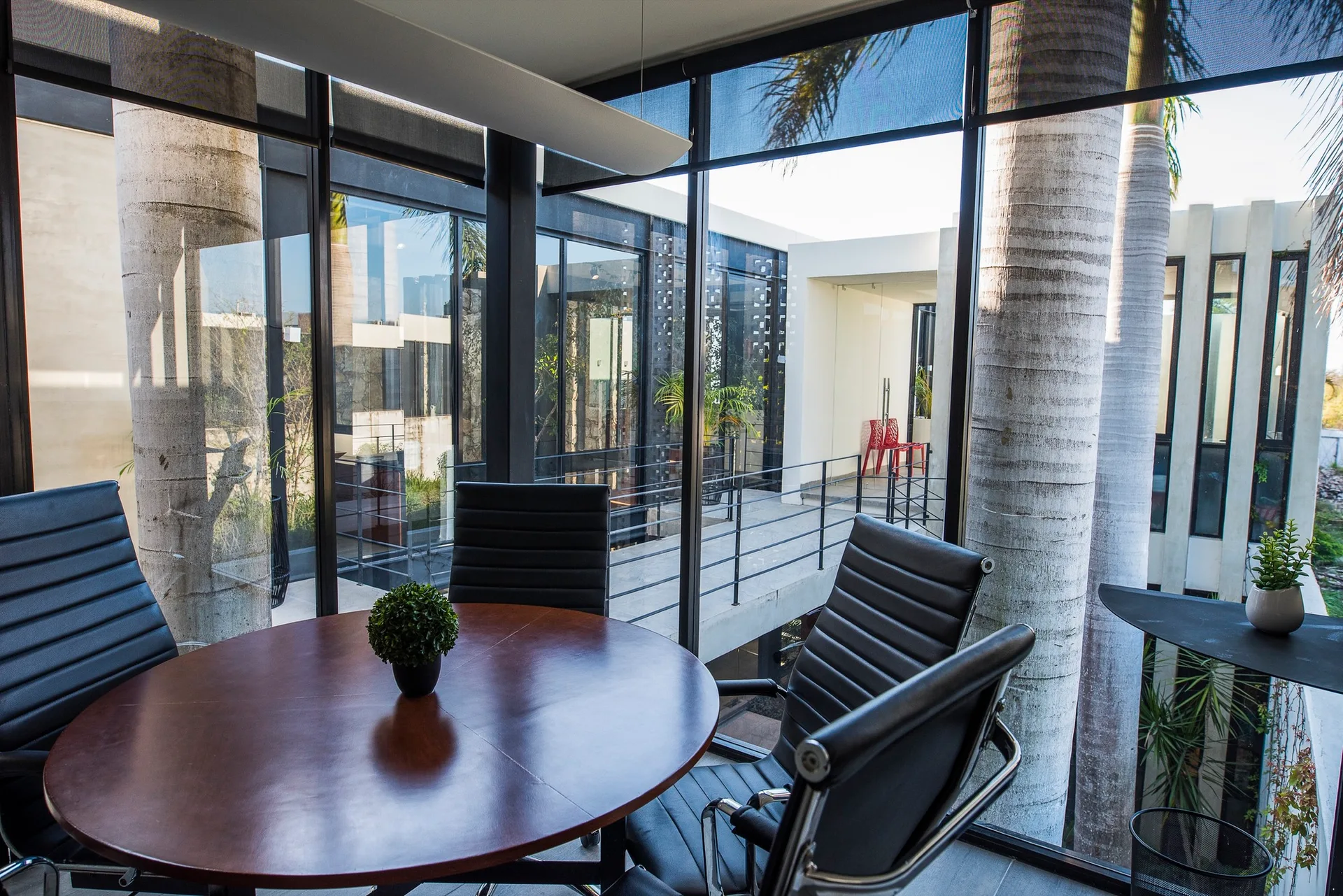 A circular wooden conference table surrounded by black ribbed office chairs is enclosed by large floor-to-ceiling glass windows showing an outdoor courtyard.